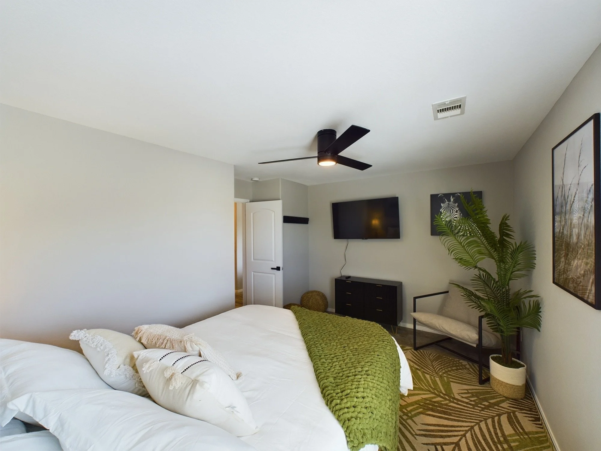 A bedroom with a white bed, decorative pillows, a green throw blanket, a black dresser, a wall-mounted TV, a large framed picture, a potted plant, a ceiling fan, and a patterned rug.