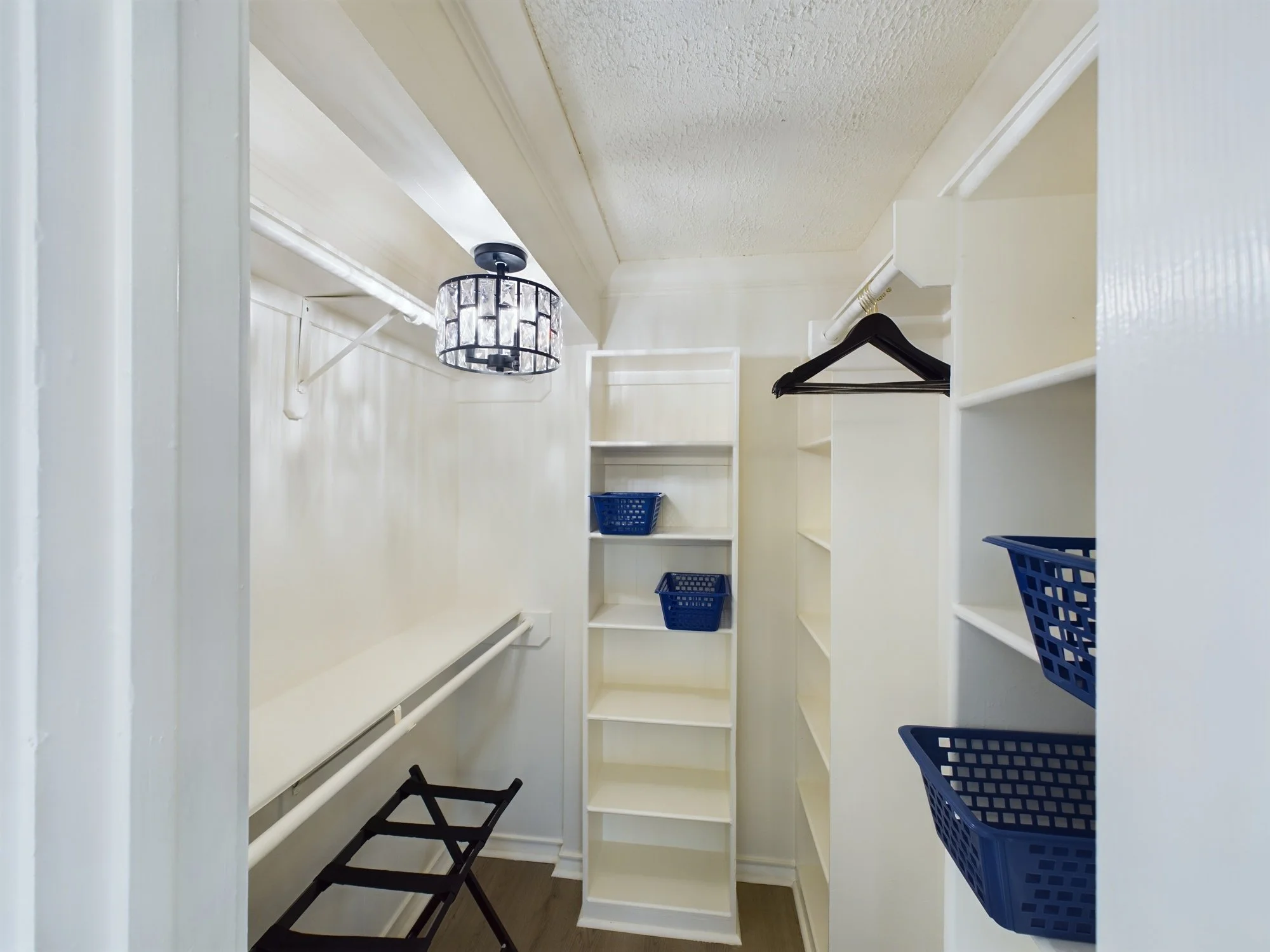 Empty walk-in closet with white shelves, hanging rod, black and blue baskets, and a ceiling light fixture.