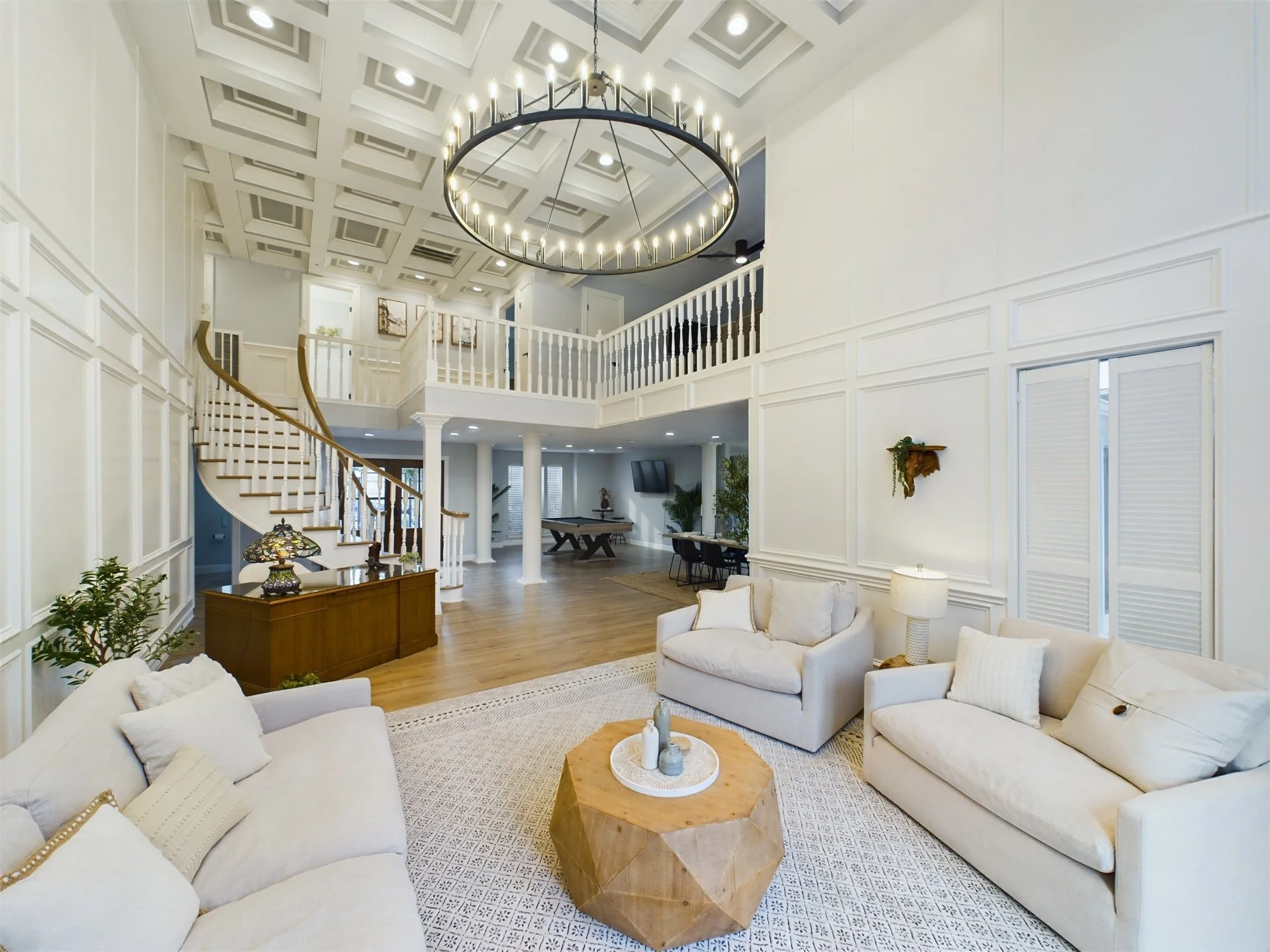 Spacious living room with white sofas, a wooden coffee table, and a patterned rug. The room features high ceilings with decorative panels, a large circular chandelier, and walls with paneled molding. There are also plants and wall art visible in the 