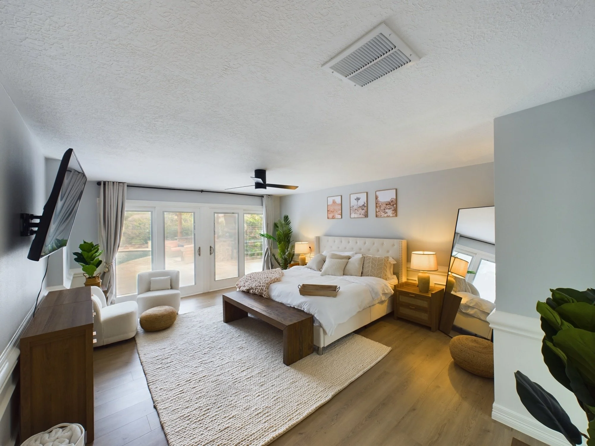 A cozy bedroom with a large bed, nightstands, and lamps, with a ceiling vent and a flat-screen TV on the wall, sunlight coming through sliding glass doors, and framed pictures of desert landscapes on the wall.