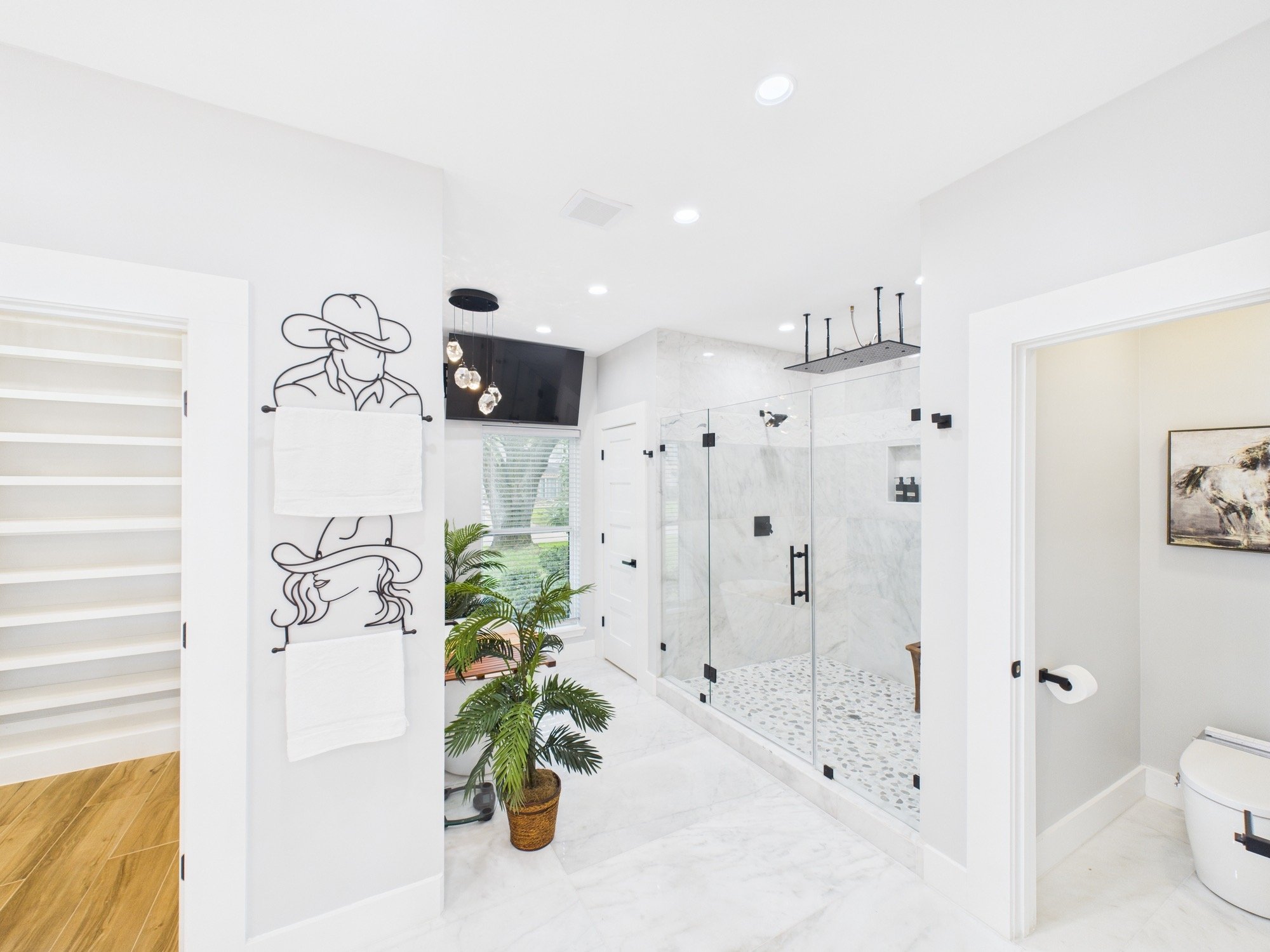 Modern bathroom with a glass shower enclosure, marble walls, and ceiling lights. There is a leafy plant in a basket, a towel rack, and artwork on the wall.