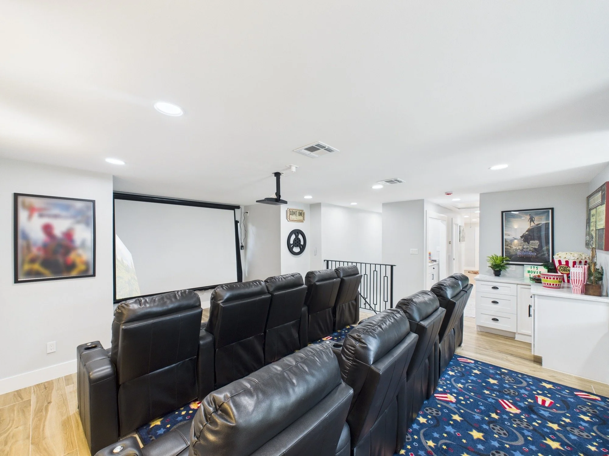 Large room with multiple rows of black leather recliner chairs, a white cabinet with popcorn and snacks, framed posters on the walls, a projector screen, and a ceiling-mounted projector, suggesting a home theater or entertainment area.