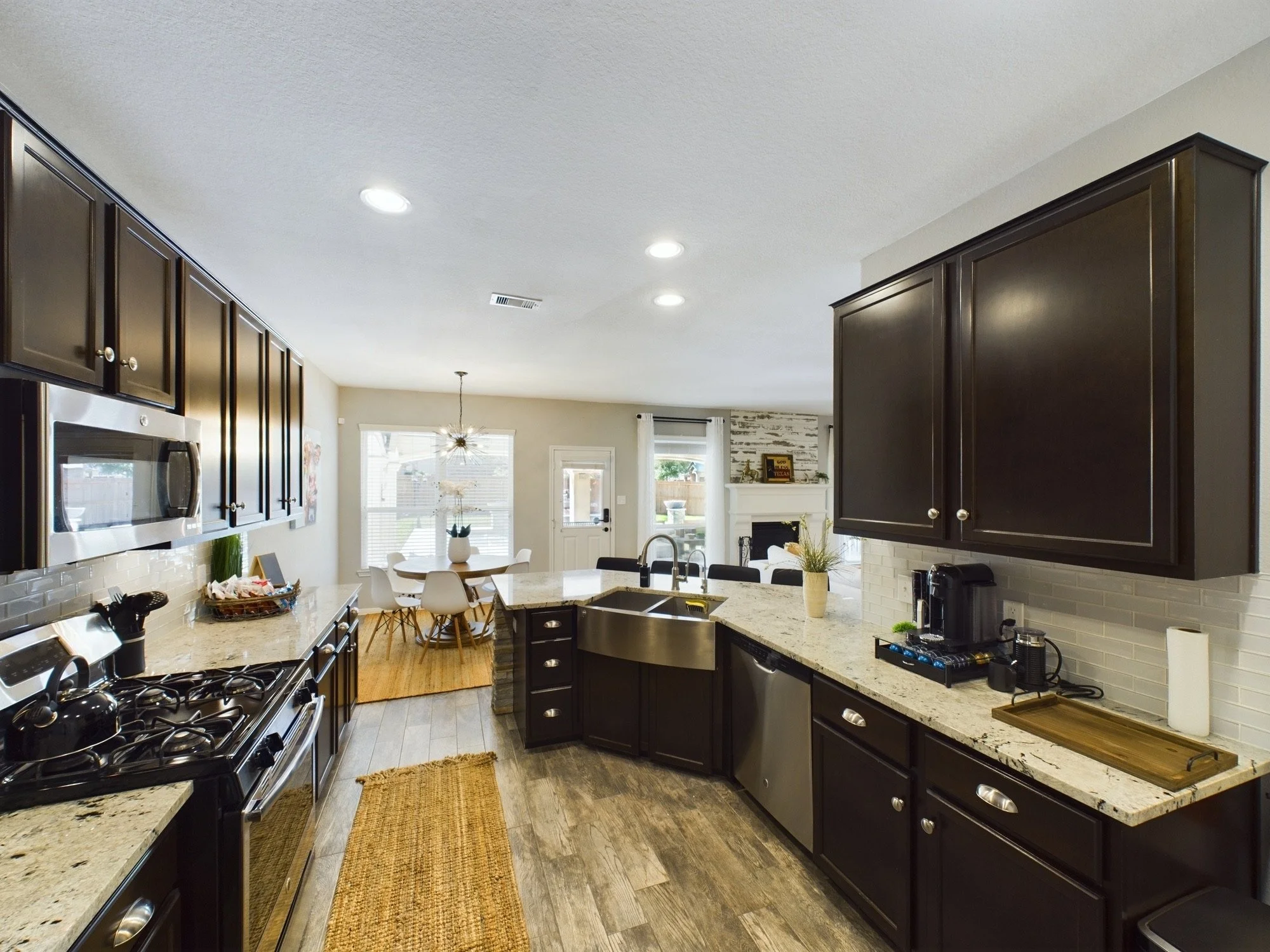 View of a modern kitchen with dark cabinets, granite countertops, stainless steel appliances, and a small island with a sink. In the background, a dining area with a round table and chairs near large windows and a living space with a fireplace and sl