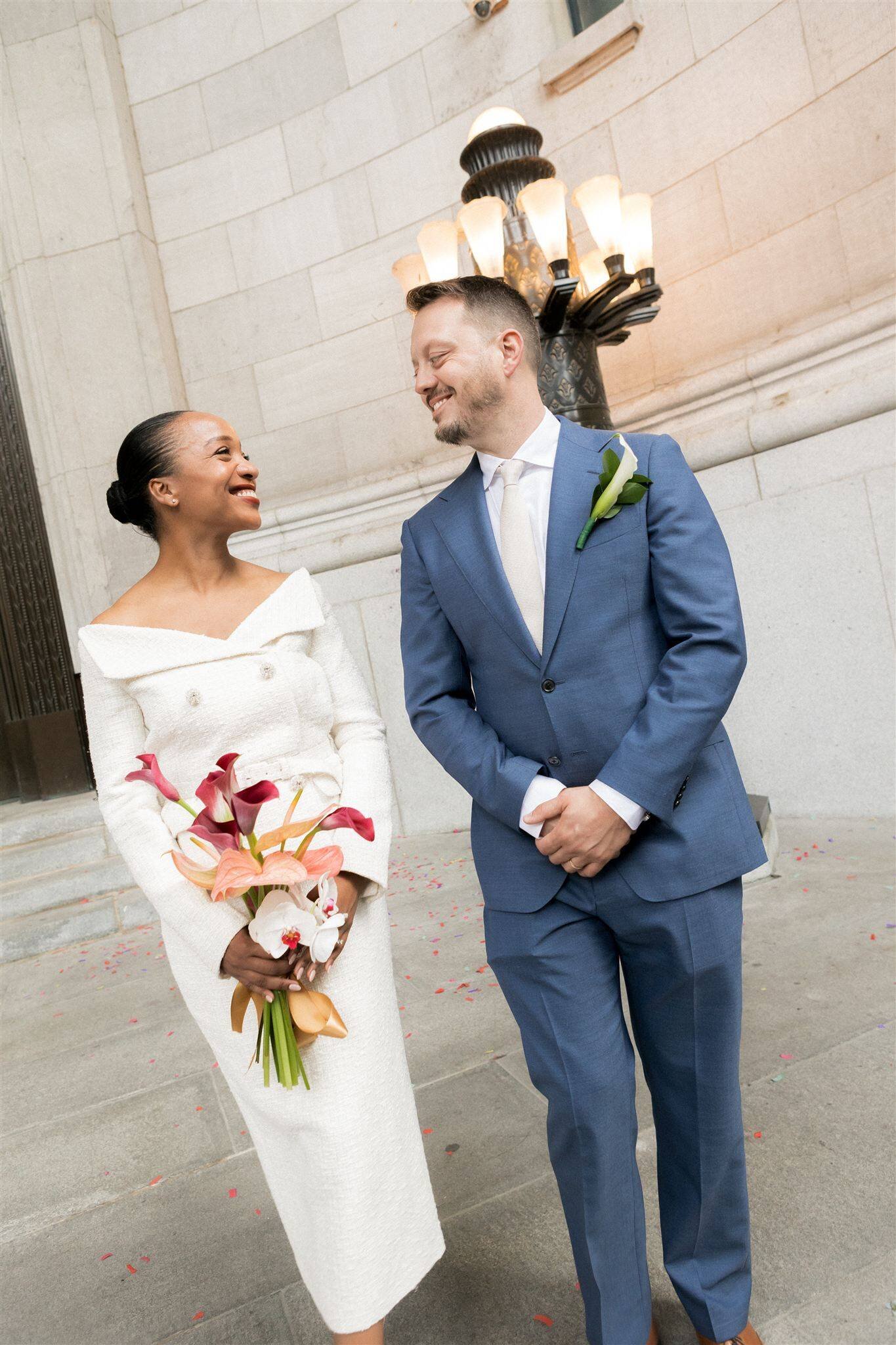A woman in a white dress holding a bouquet of pink and white flowers, standing next to a man in a blue suit with a white tie and a white calla lily pinned to his lapel, smiling and looking at each other outside a stone building with a large lighting fixture in the background.