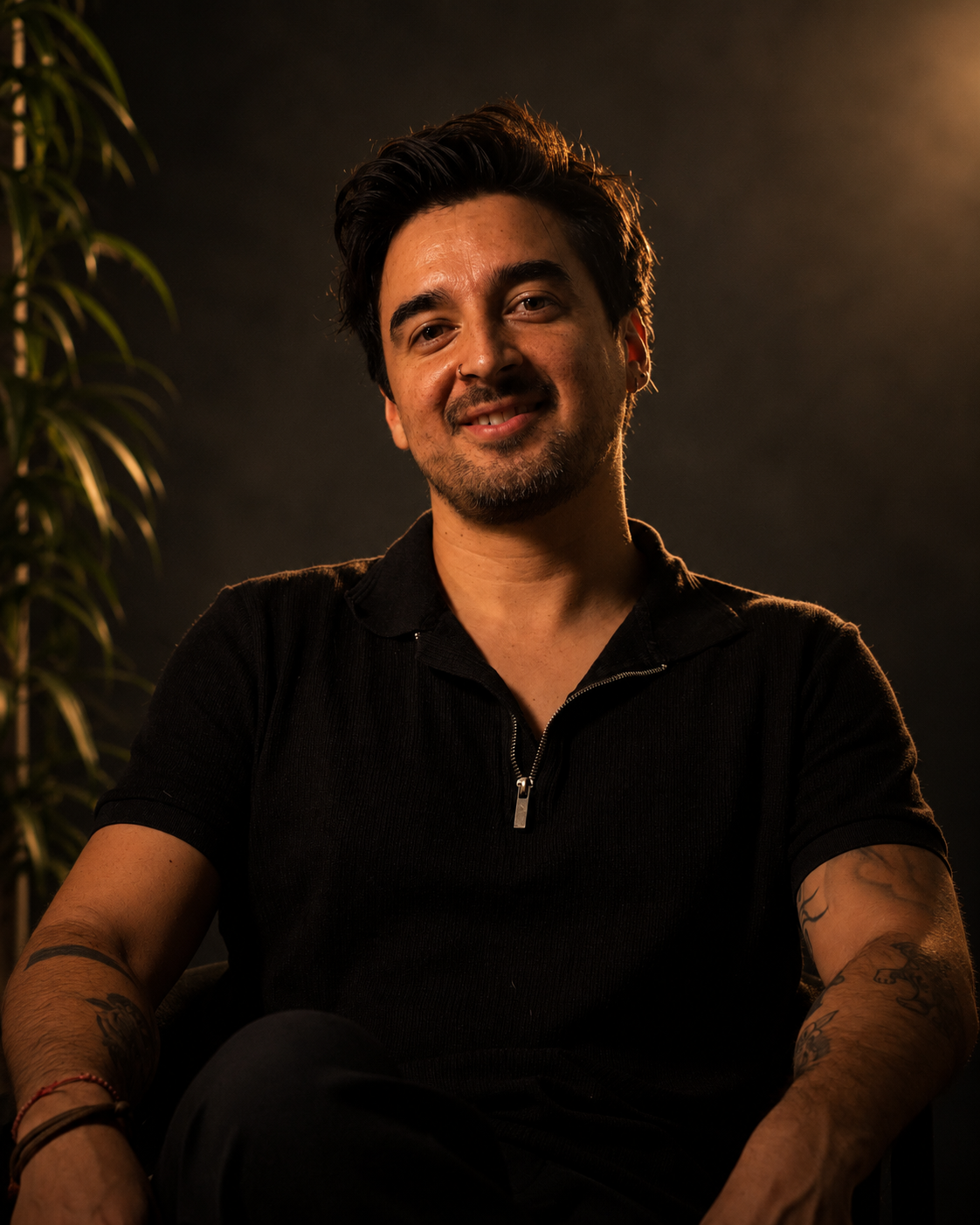 Joven hombre con barba y tatuajes, sonriendo, en un entorno con iluminación cálida y fondo oscuro.