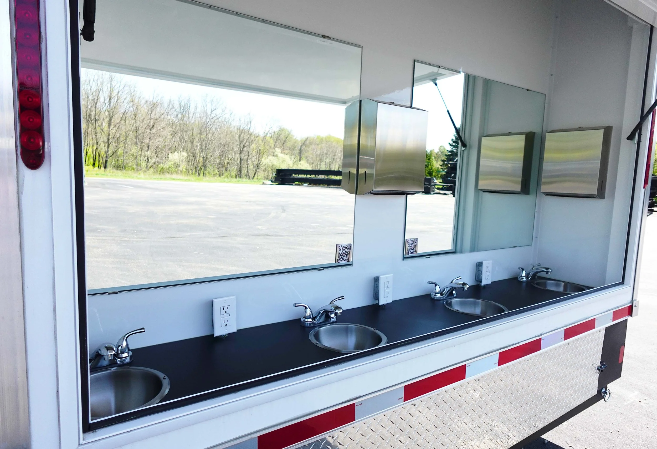 A mobile washing station with three sinks, mirrors, electrical outlets, and soap dispensers inside a service trailer, with an outdoor view of trees and a parking lot.