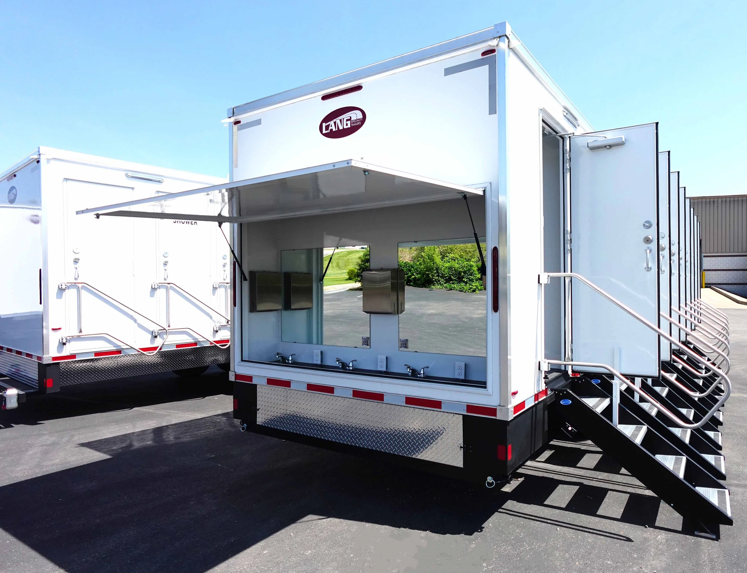 A white mobile trailer with stairs and a ramp, featuring a large window, outdoor sinks, and a logo that reads "LANG Specialty Trailers."