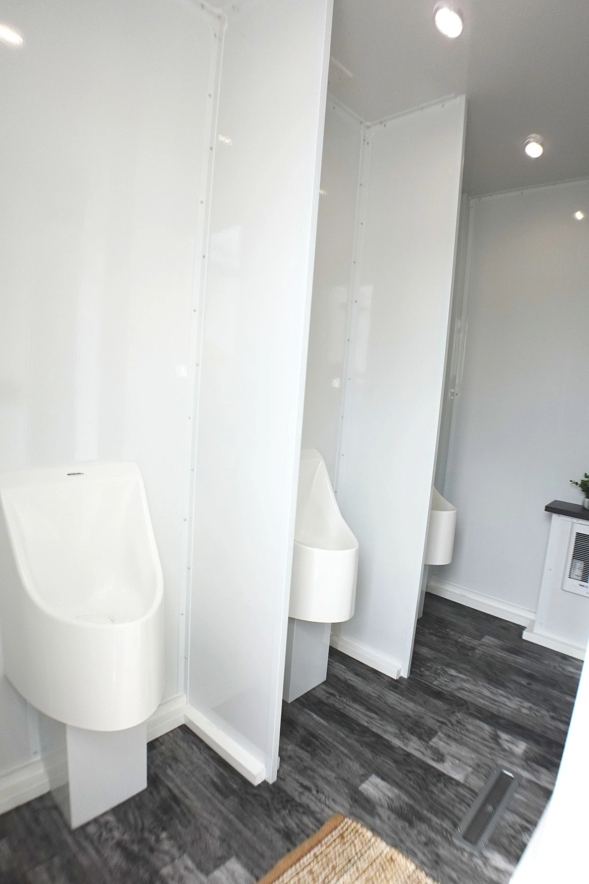 Public restroom with three white urinals separated by white partitions, a white wall with a vent, and dark wood-look flooring.