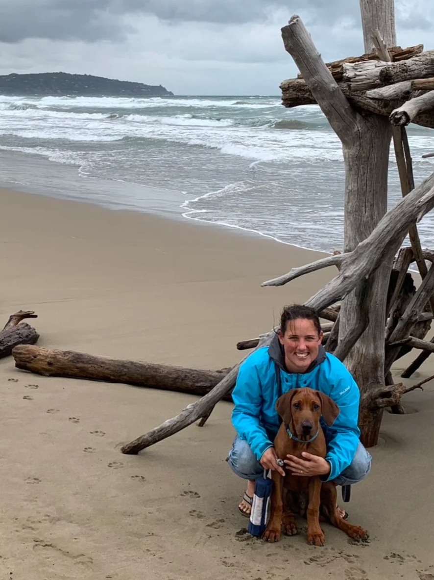 Hundetrainerin mit ihrem Hund an einem stürmischen Strand, mit Treibholz im Sand und Wellengang im Hintergrund.