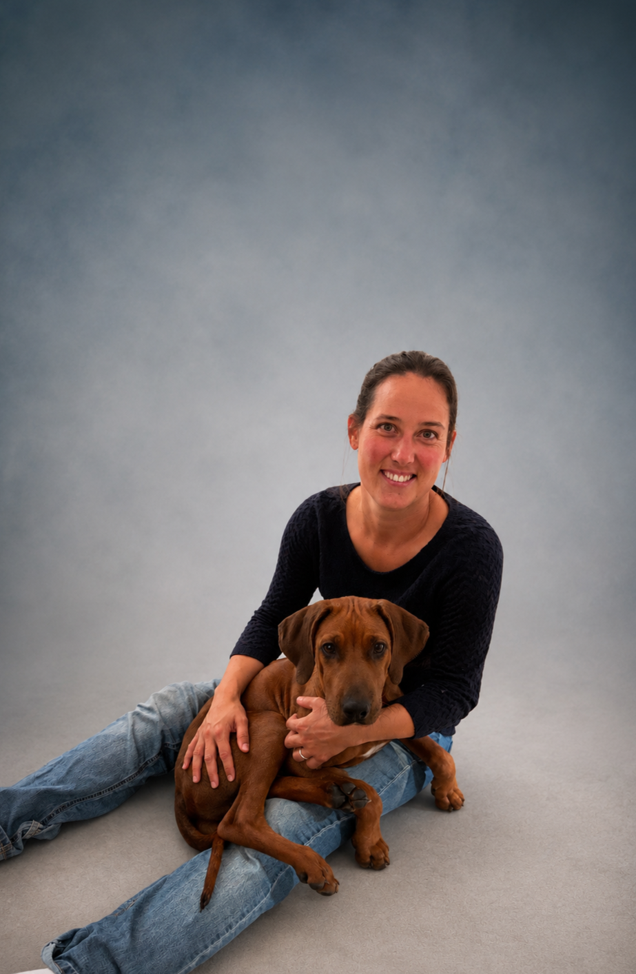 Eine lächelnde Frau sitzt auf dem Boden, ein Rhodesian Ridgeback Junghund liegt zwischen ihren Beinen und auf ihrem Schoß.