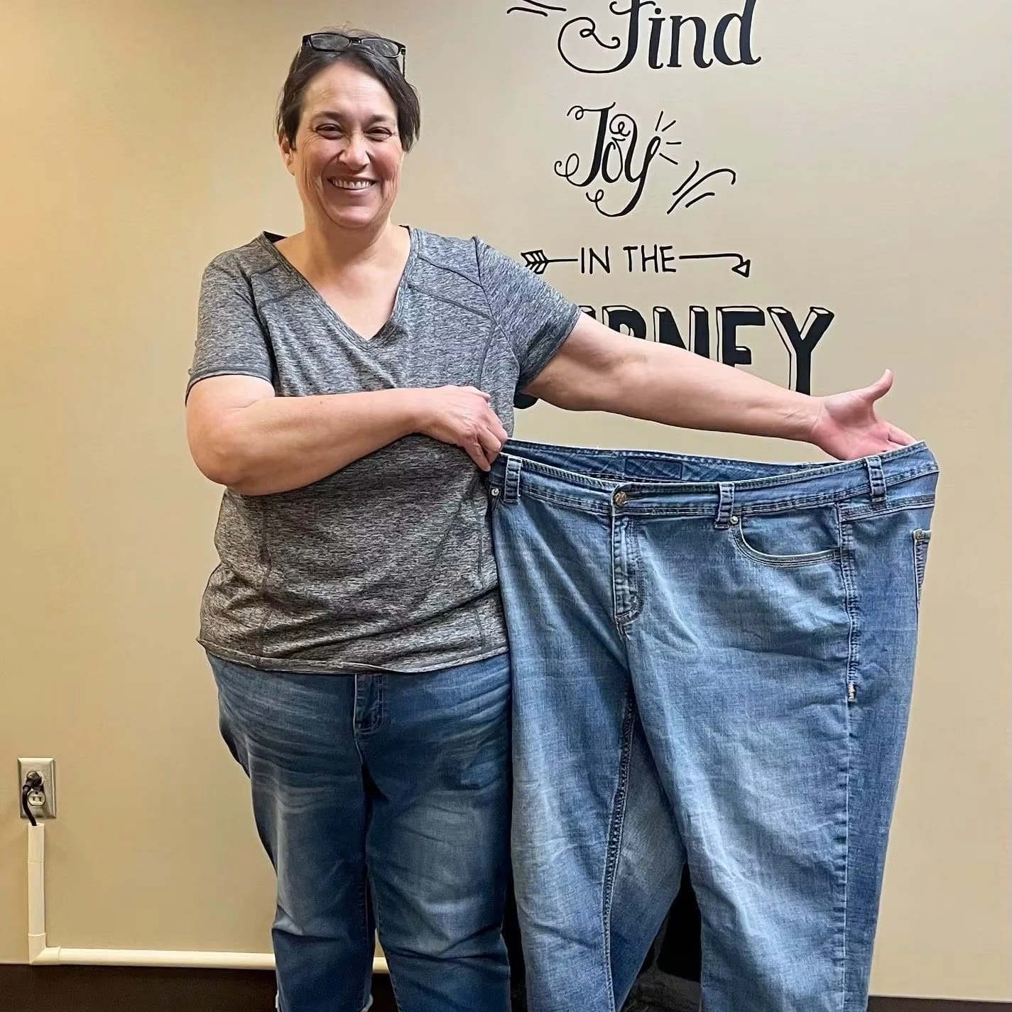 Woman smiling and holding a pair of large jeans, standing in front of a wall with inspirational quote.