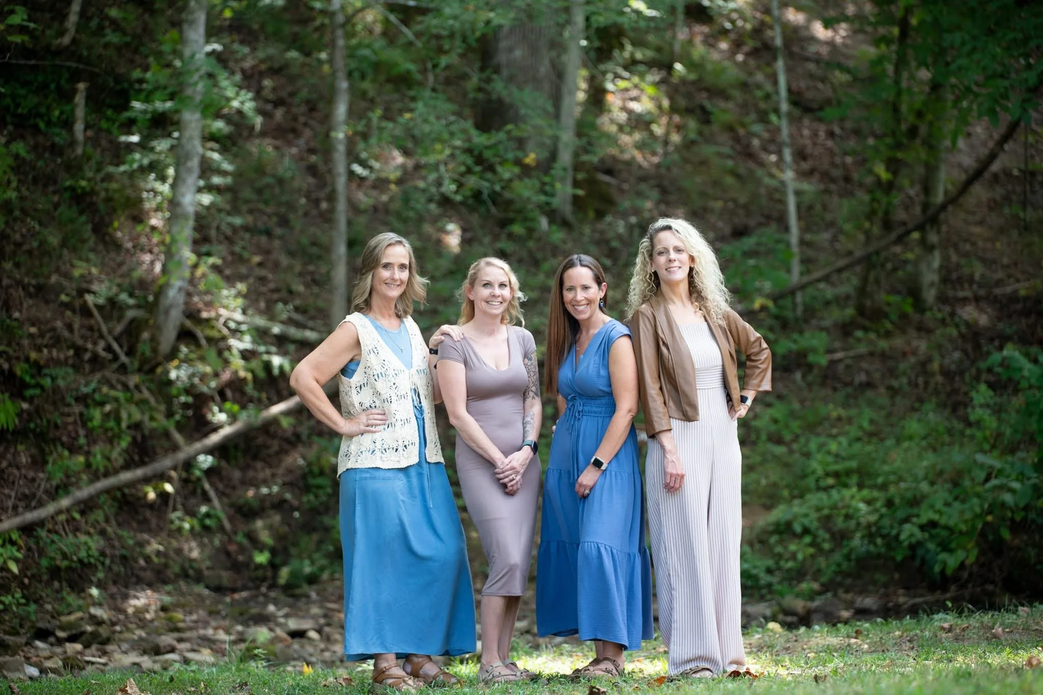 Four women standing outdoors in front of a wooded area, smiling at the camera, with a mix of casual and semi-formal clothing.