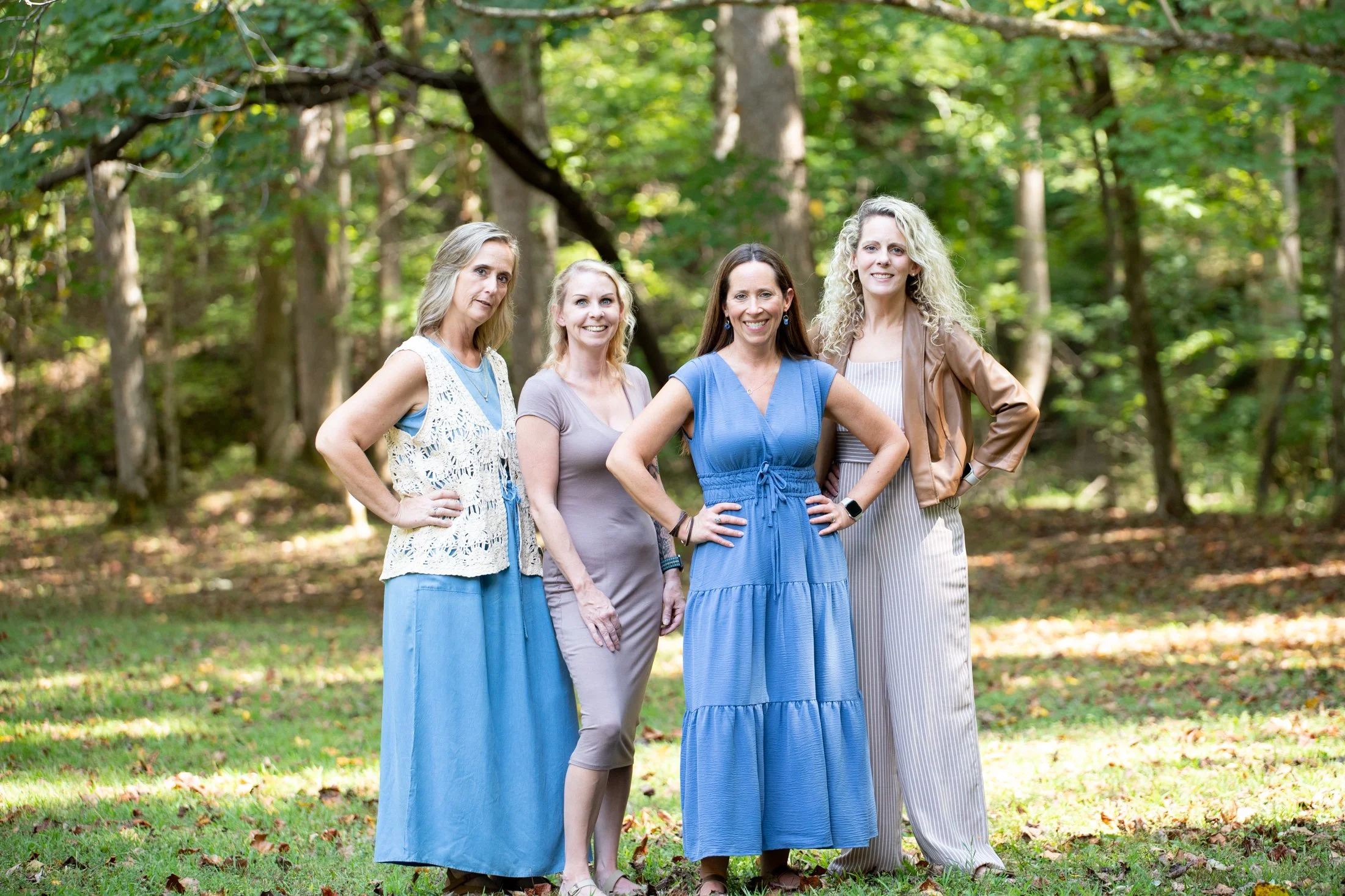Four women standing outdoors in a wooded area with trees in the background, posing for a photo.