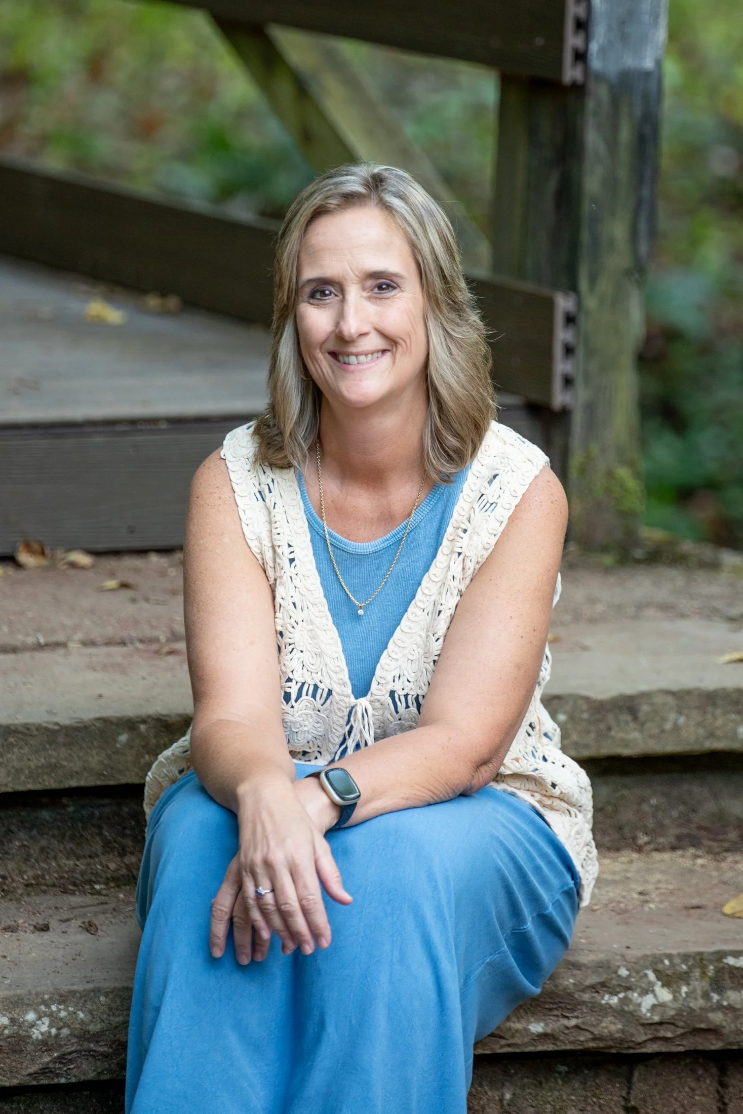 A woman with shoulder-length blonde hair, wearing a blue dress and a white crocheted vest, sitting on wooden steps outdoors, smiling at the camera, with a blurred green background.