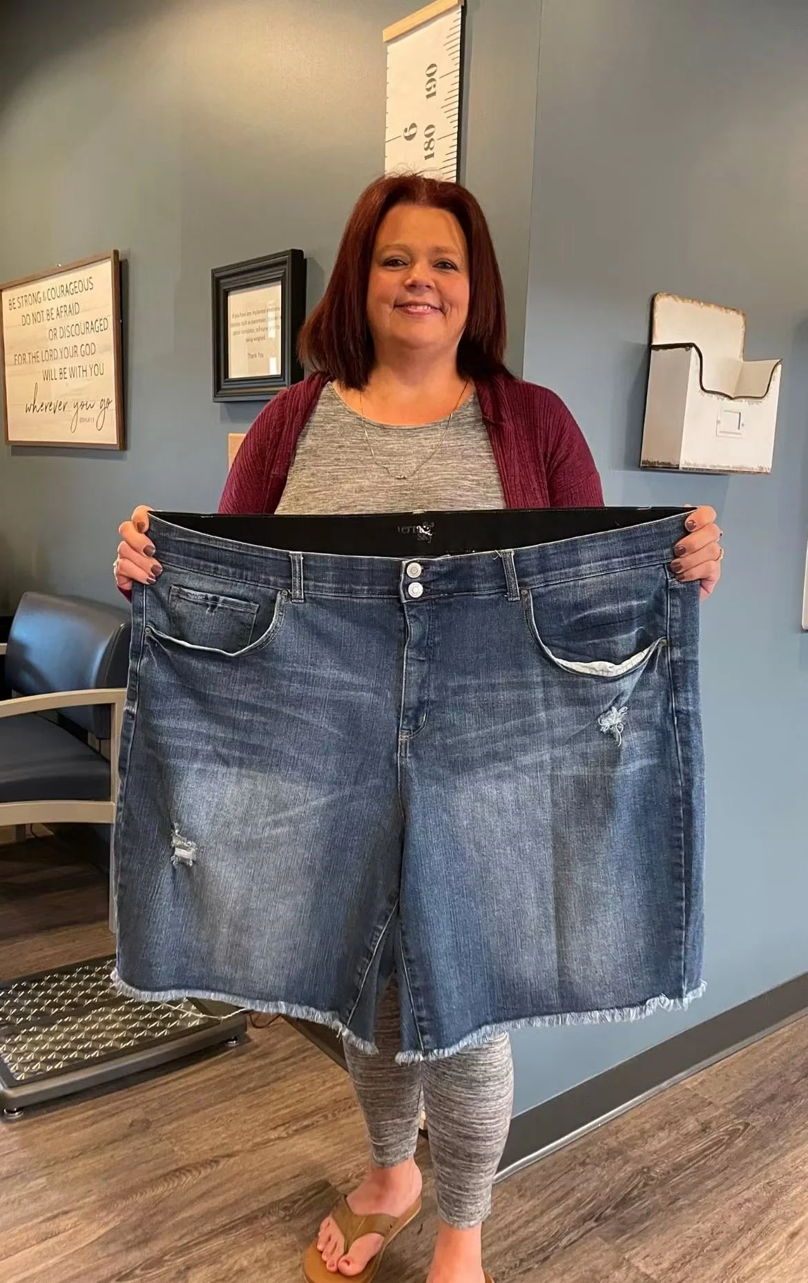 A woman with reddish-brown hair, wearing a gray shirt, gray leggings, and tan sandals, holding large blue jeans in front of her in a room with blue walls and framed artwork.