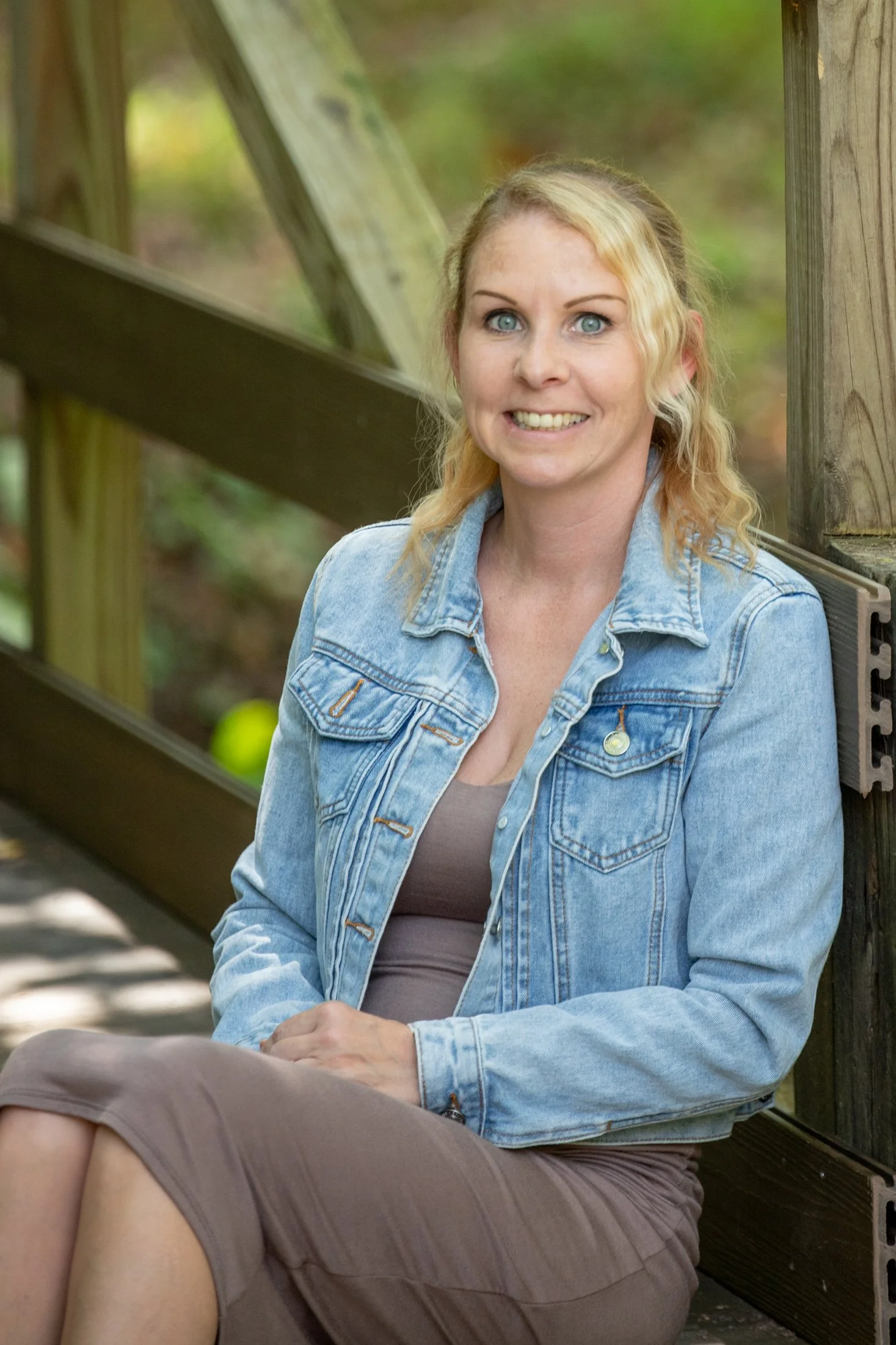 A woman with blonde, curly hair and blue eyes smiling while sitting on a wooden bench outdoors. She is wearing a light blue denim jacket and a brown top and skirt. The background is blurred greenery.