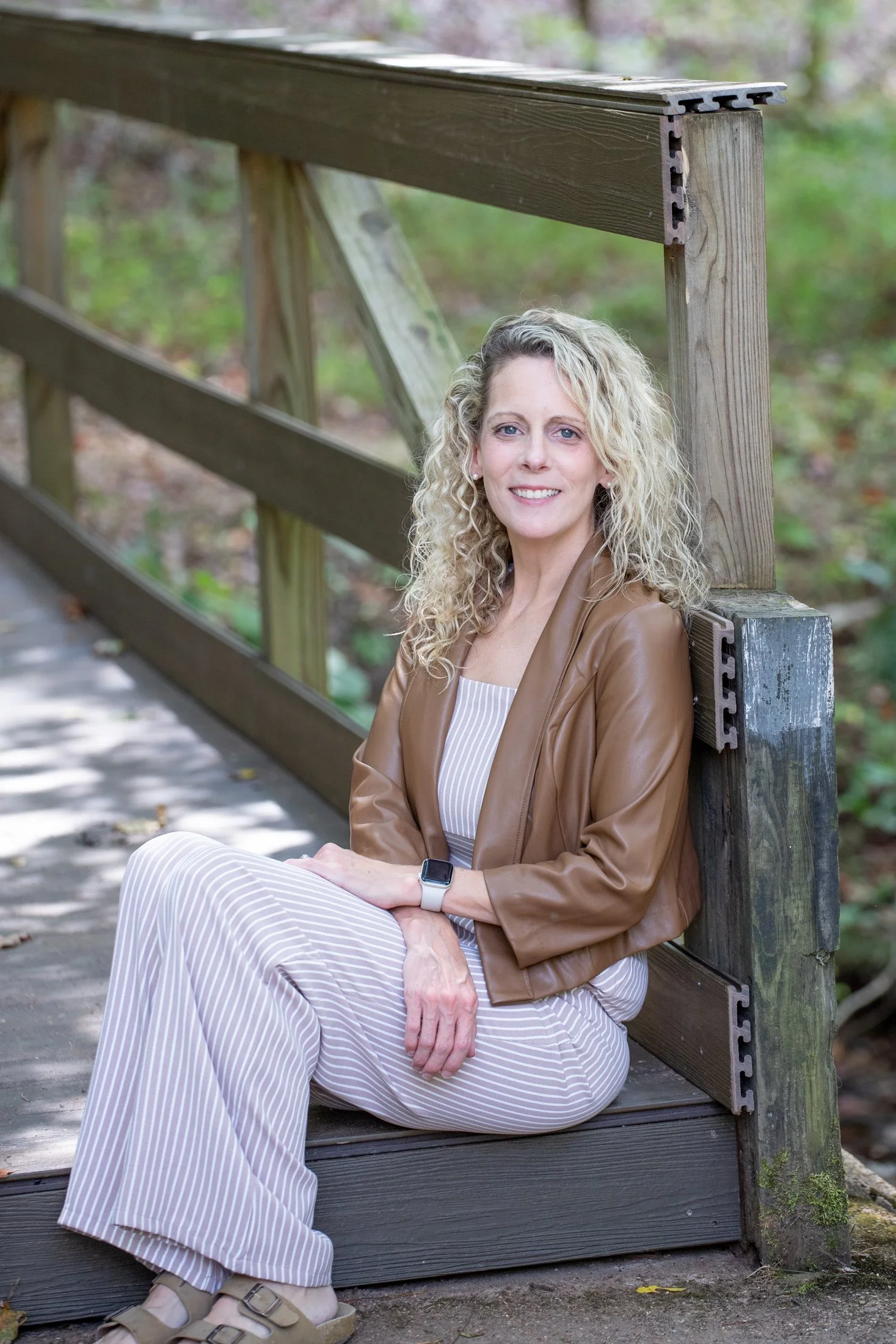 A woman sitting on a wooden park bench, wearing a brown leather jacket, striped pants, and sandals, smiling at the camera with a background of blurred green trees.