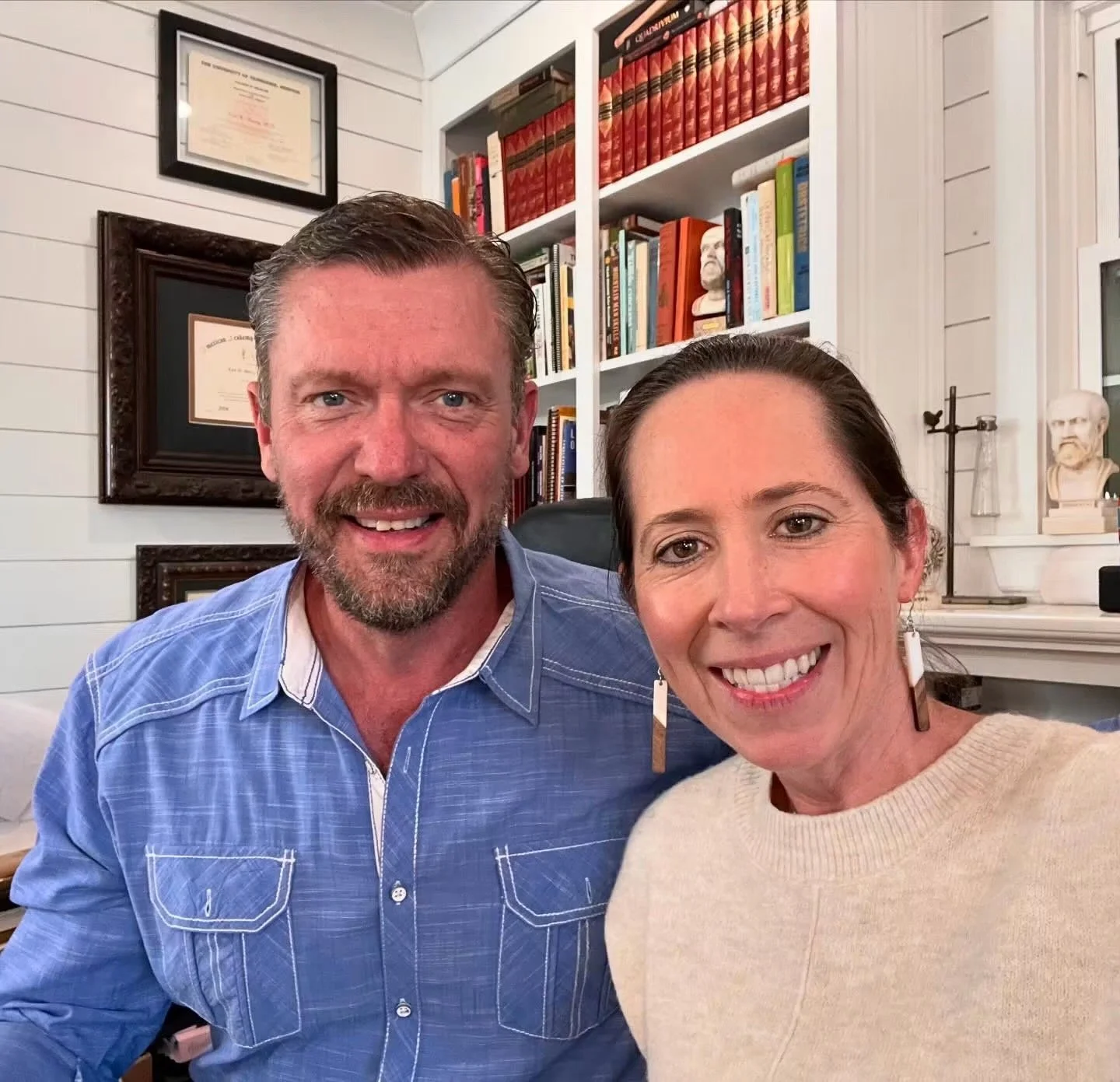 A man with a beard and a woman with dark hair smiling for a selfie in a room with white shiplap walls, a bookshelf filled with books, framed certificates, and bust sculptures in the background.