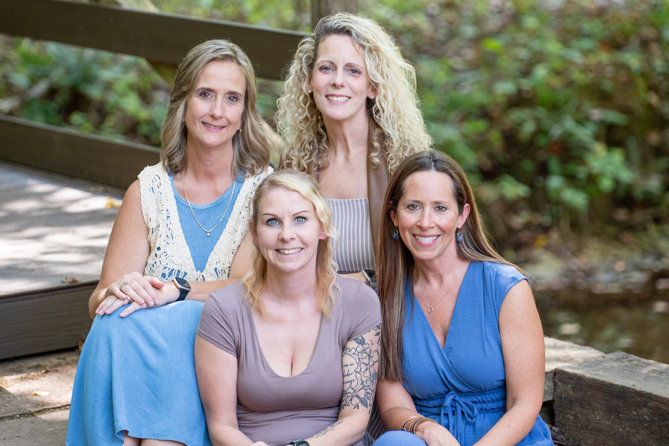 Five women with varying hair colors and styles sitting outdoors on wooden steps surrounded by green foliage, smiling at the camera.