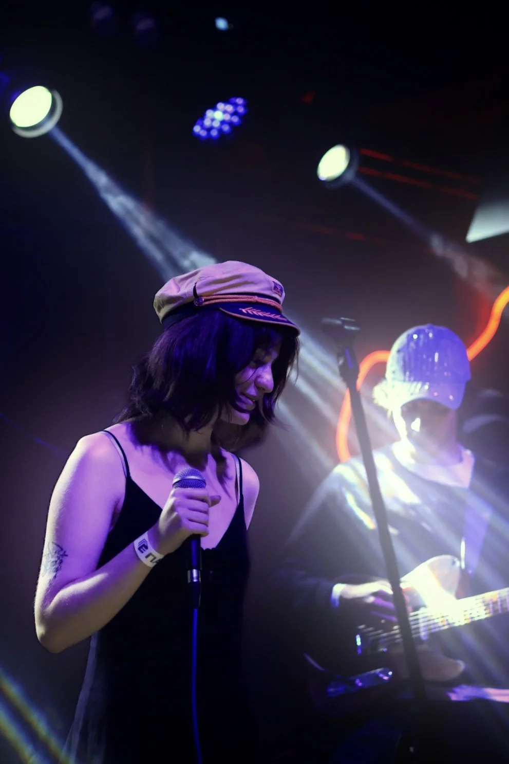 A woman in a black dress and a tan hat singing into a microphone on stage while a guitarist with a glittery bucket hat plays behind her, illuminated by stage lights.