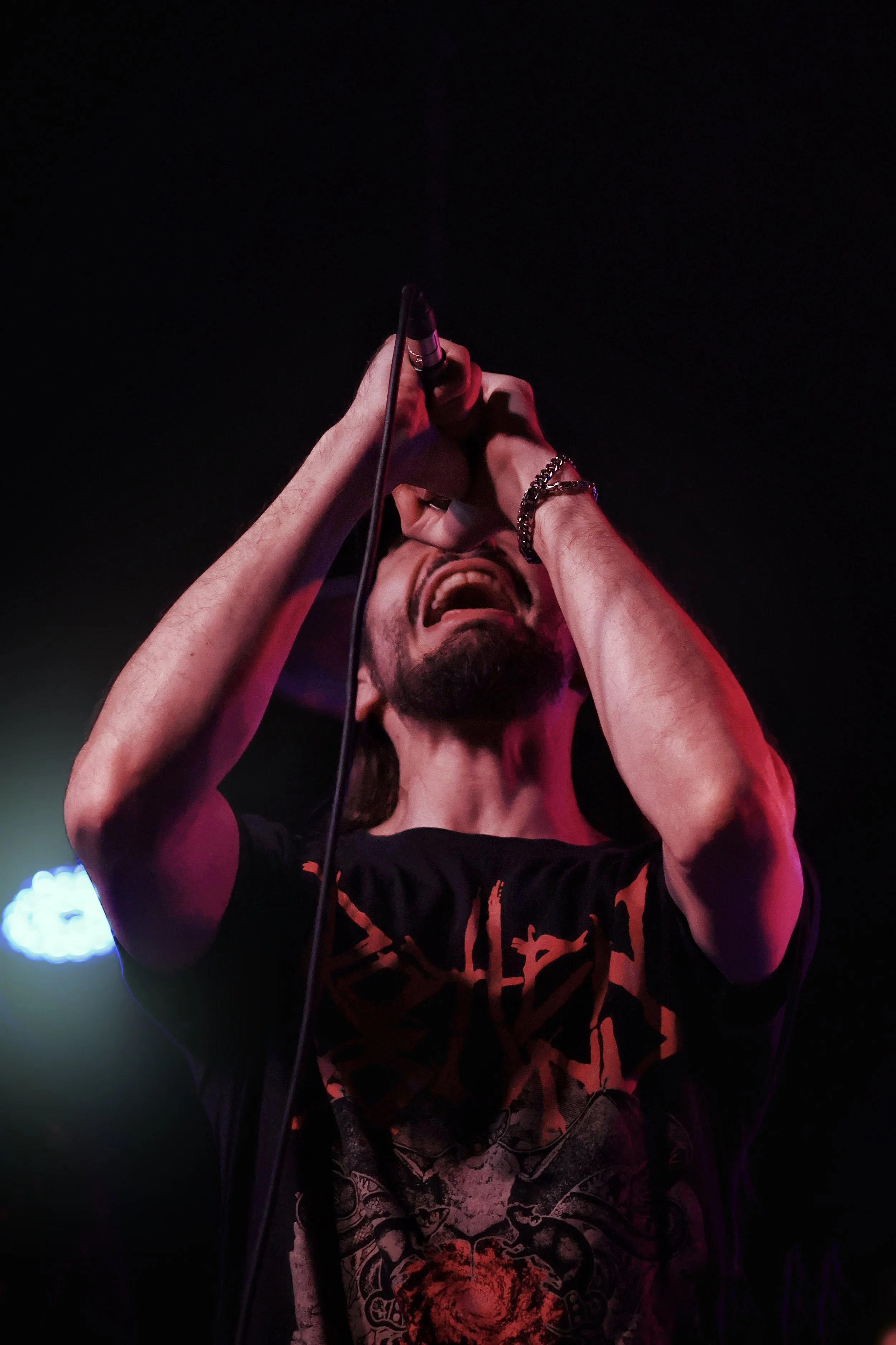 A person with long hair and a beard, wearing a black T-shirt with a graphic design, holding a microphone and performing on stage with dark background and colorful stage lighting.