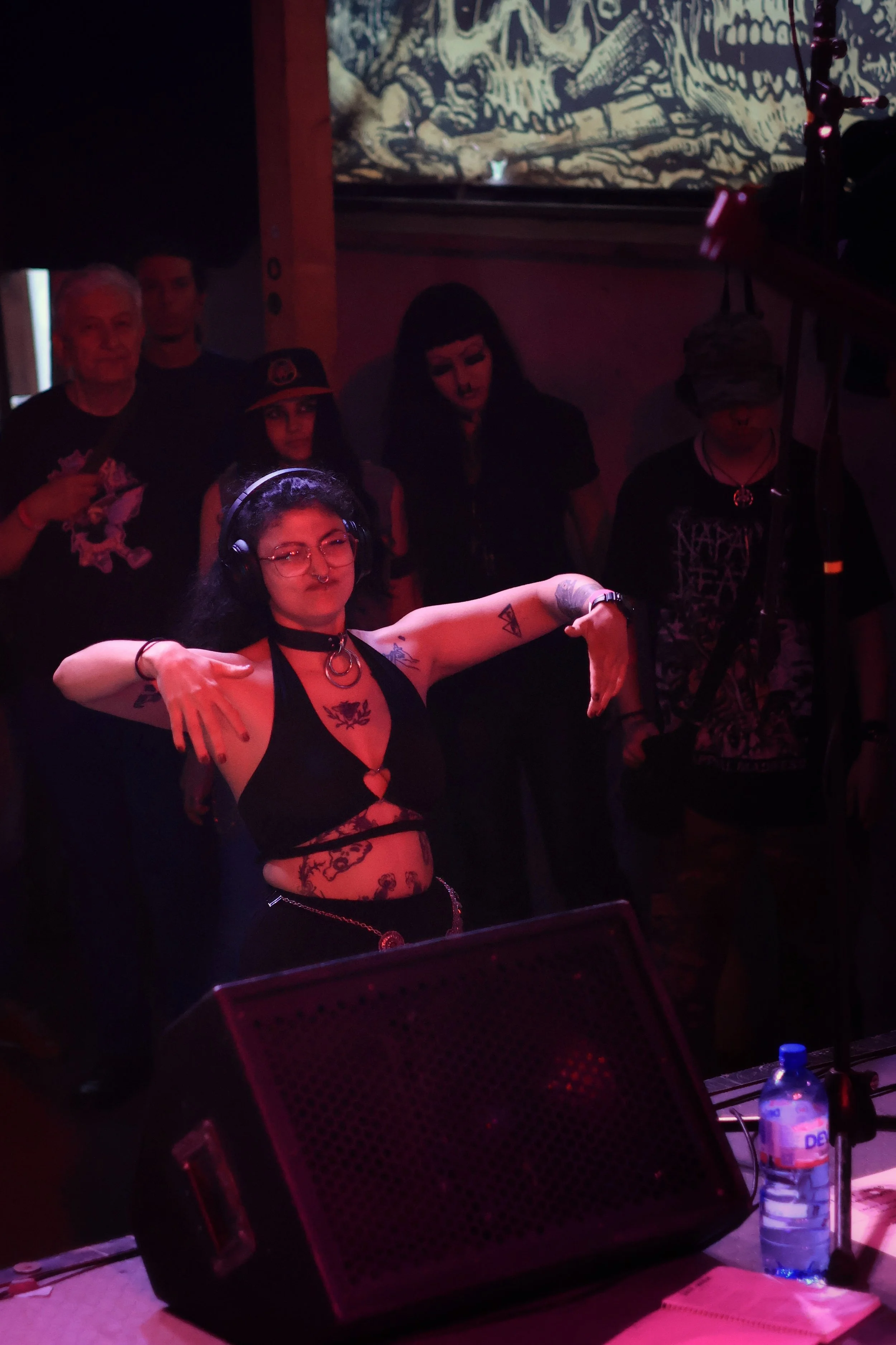 A group of five people gathered in a dimly lit room with dark ambiance, possibly a nightclub or concert setting. The central figure is a woman with dark hair, glasses, and tattoos, wearing a revealing black outfit with a choker and piercings, striking a pose with her arms out.