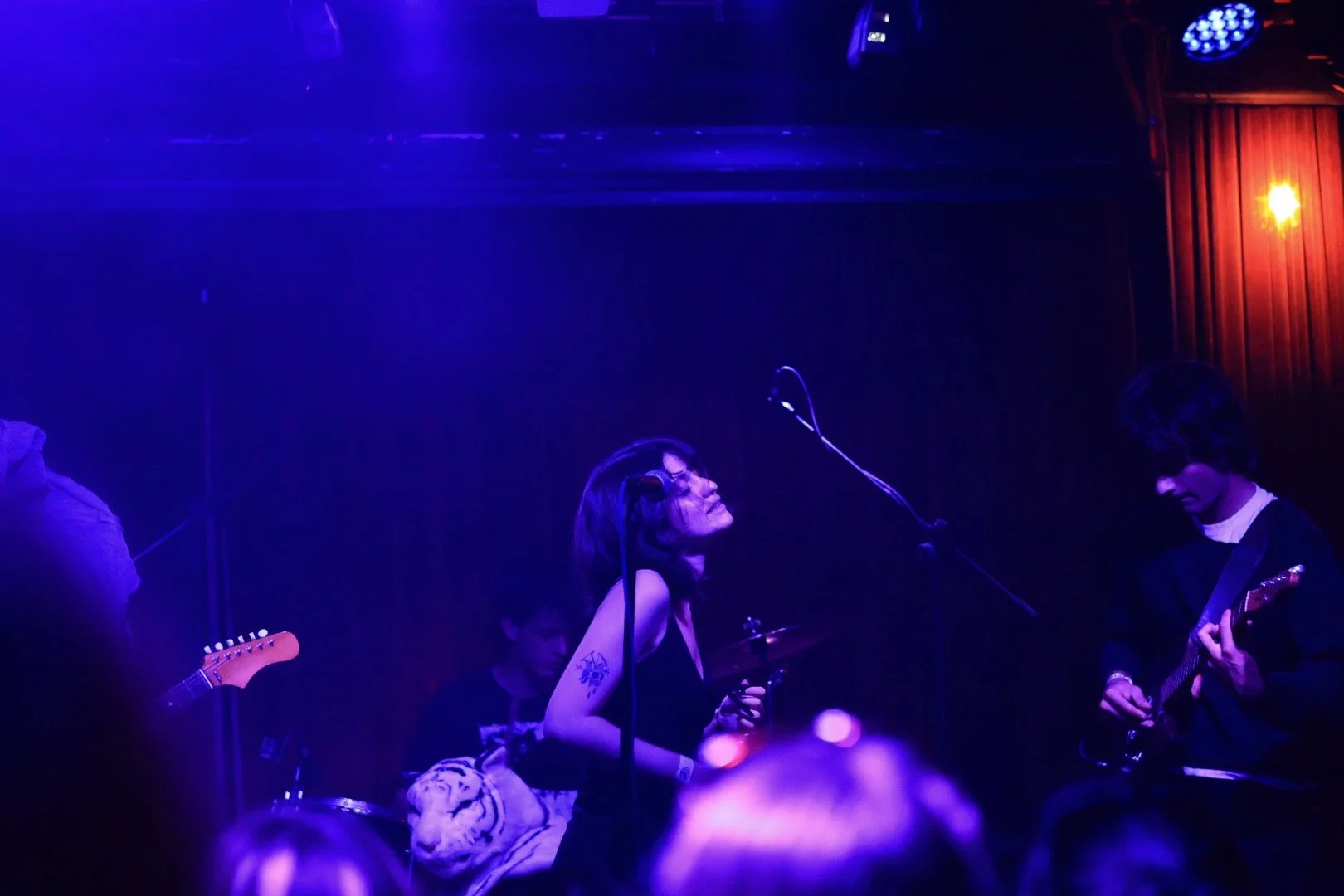 Band performing on stage in a dimly lit setting with purple and orange lighting. The lead singer, a woman with short dark hair, is smiling and singing into a microphone. To her right, a guitarist is playing, and in the background, a keyboard player i