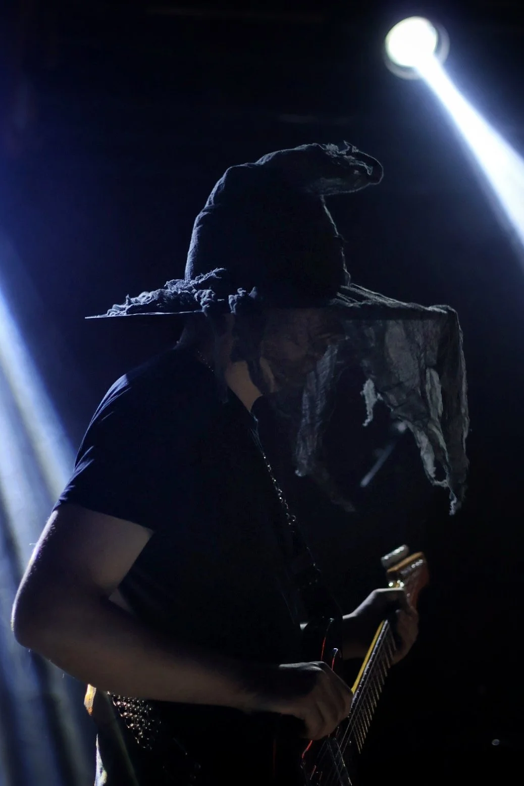 Person wearing a large witch hat and a tattered mask playing an electric guitar on stage with spotlights.
