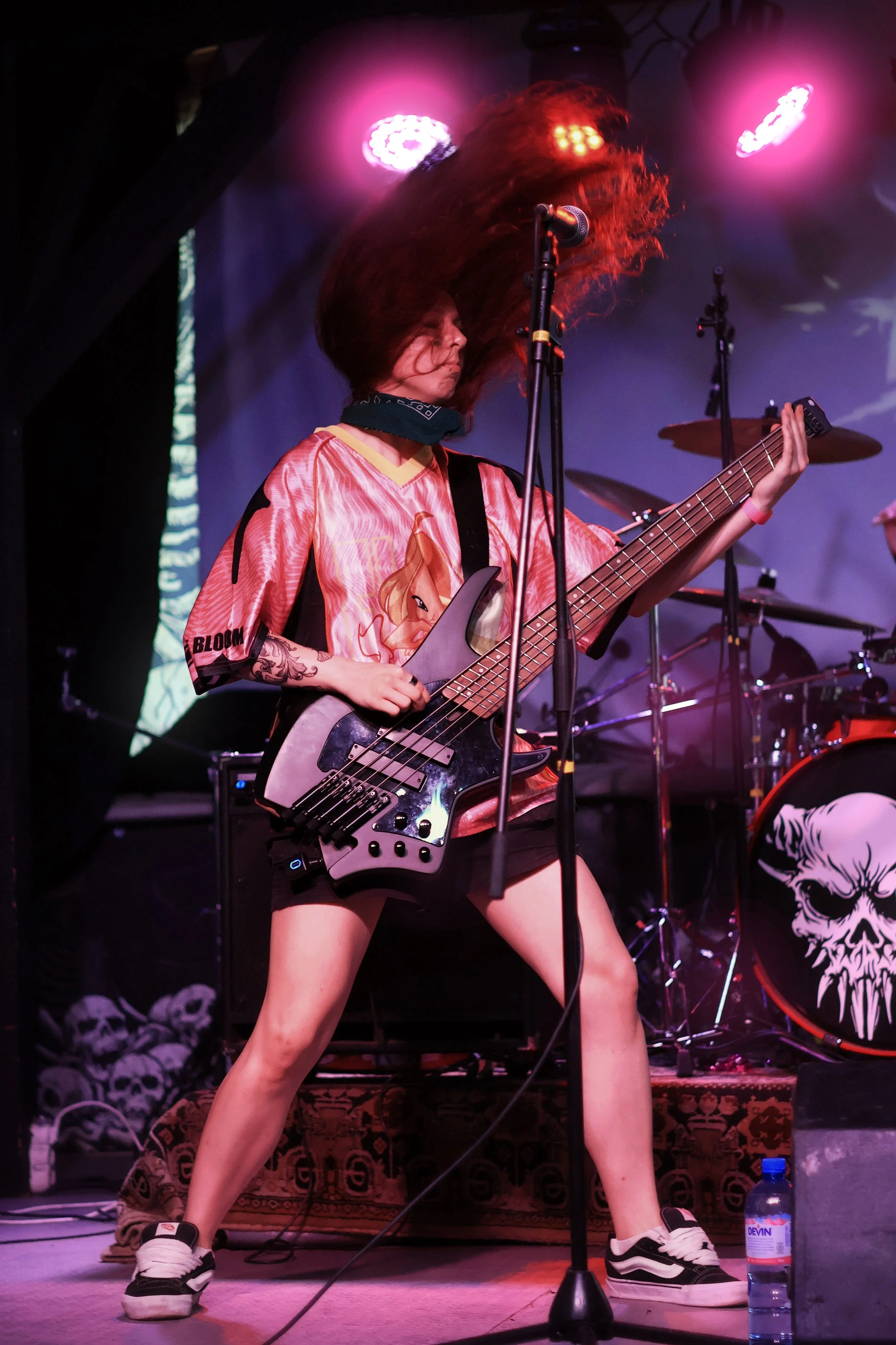 A female musician with long red hair headbanging while playing an electric bass guitar on a stage with pink and purple lights, wearing a cartoon character jersey, shorts, and sneakers.