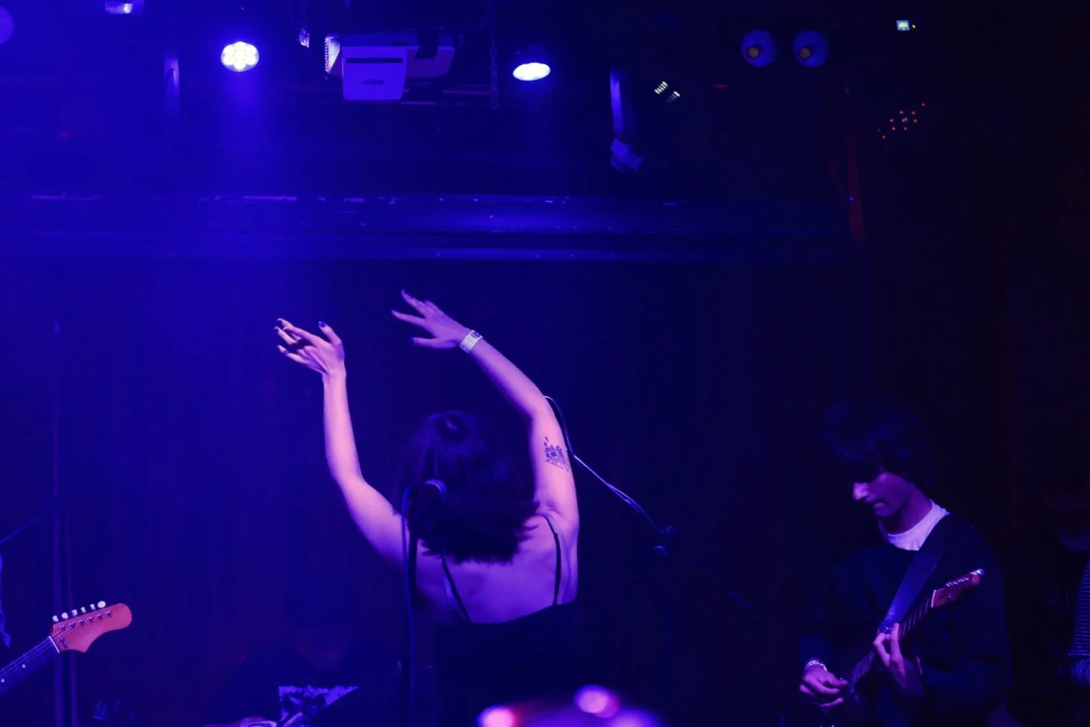 A woman with short dark hair dancing with her arms raised on stage, illuminated by blue and purple stage lights, with a male guitarist playing guitar nearby in a dark venue.