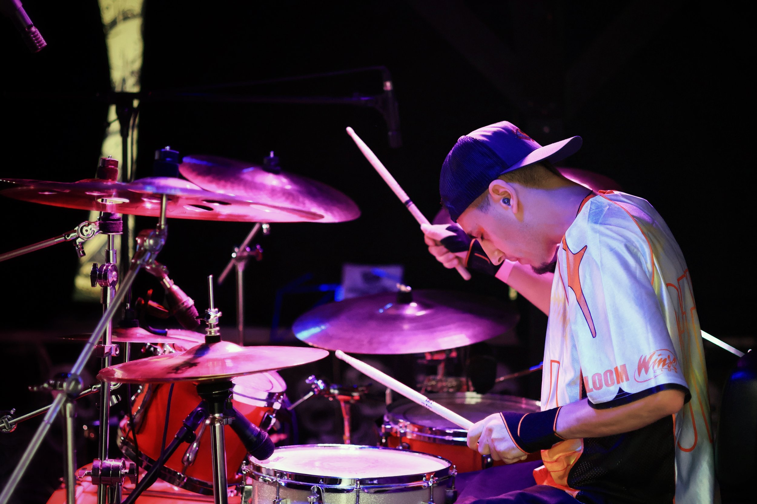 A musician wearing a baseball cap and a colorful jersey playing drums on stage with purple and pink stage lighting.