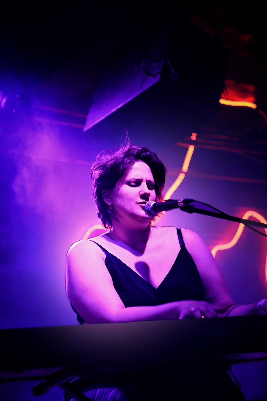 A woman with short hair singing into a microphone on stage with purple lighting and neon signs in the background.