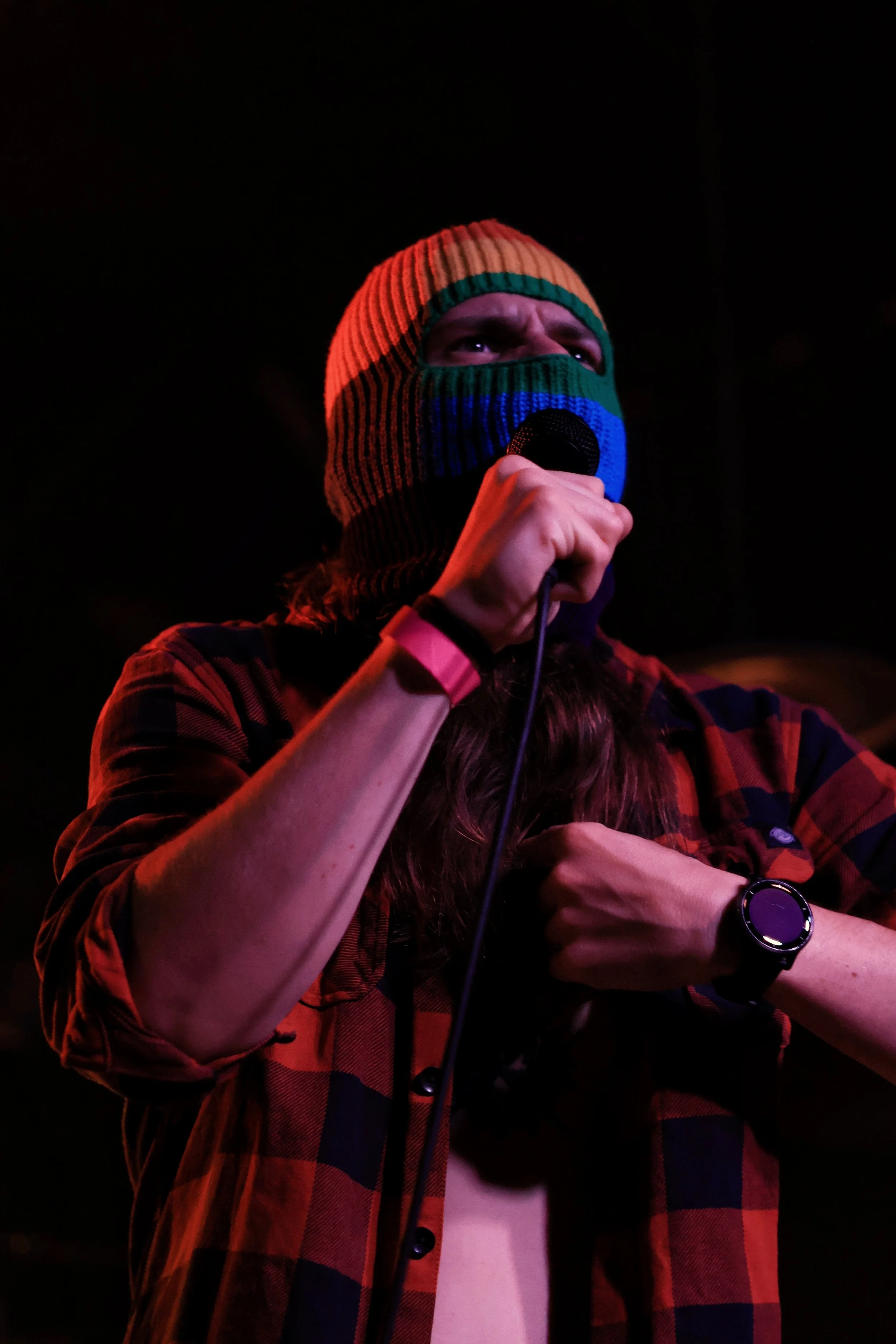 Person wearing a striped orange, green, blue, and black ski mask and a red and black flannel shirt, holding a microphone in the dark.