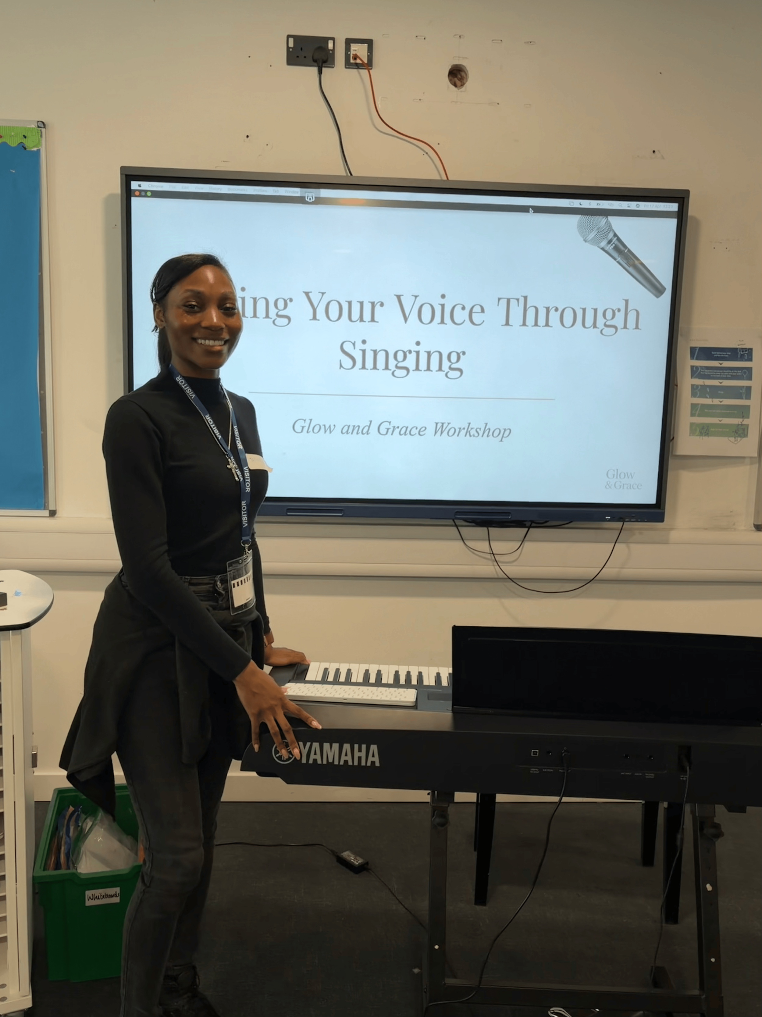 A woman smiling and playing a Yamaha keyboard in front of a large screen that displays a presentation titled "Using Your Voice Through Singing" at the Glow and Grace Workshop.