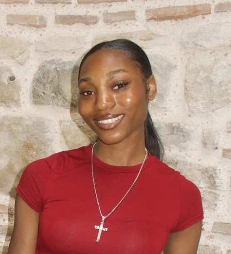 A woman with dark hair pulled back, smiling, wearing a red shirt and a silver cross necklace, standing against a brick wall.