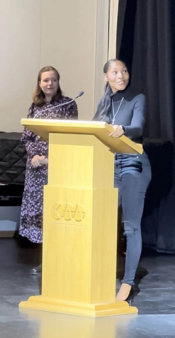 Two young women standing behind a yellow podium with a microphone, during a speech or presentation, with one woman speaking and the other listening on stage.