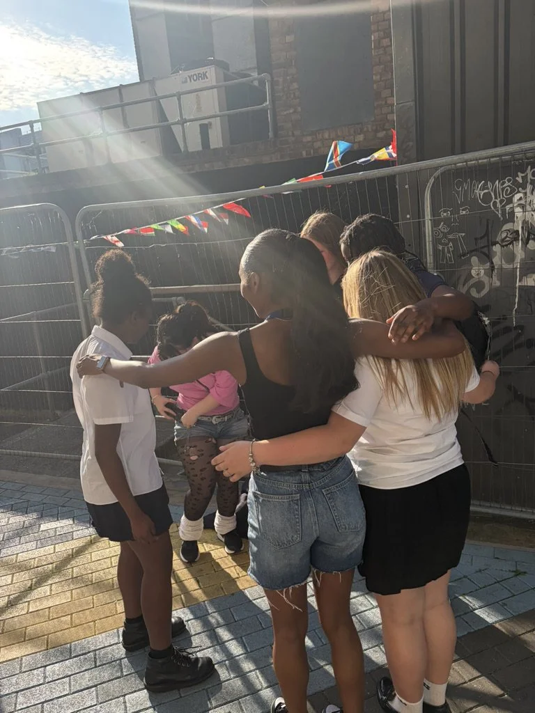 Group of children and teenagers hugging and standing together outdoors near a metal fence with graffiti, with ragged flags and a brick building in the background.