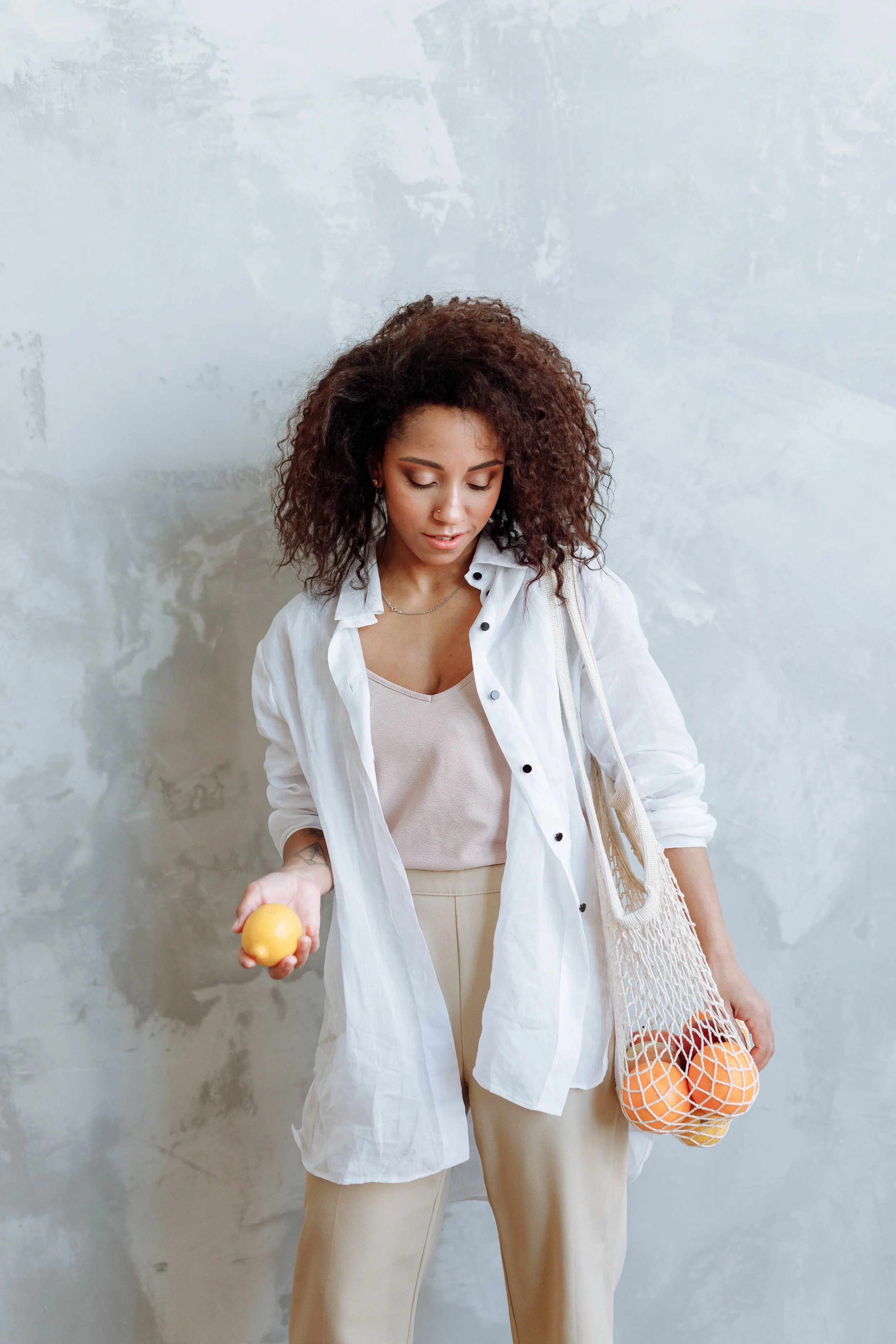 A woman with curly hair looking down and holding a yellow lemon in her right hand, while holding a net bag filled with oranges in her left hand, against a light gray textured wall.