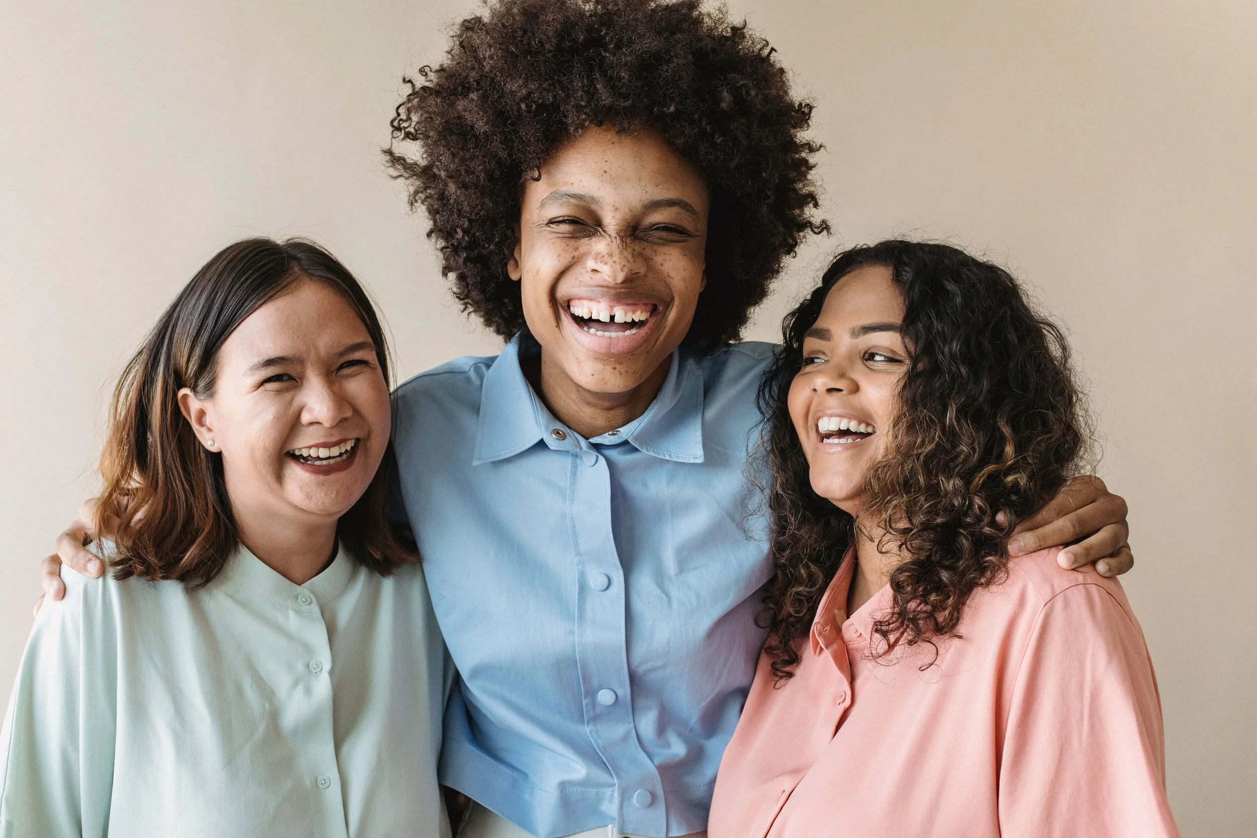 Three women standing together smiling and laughing, embracing each other in front of a beige wall.