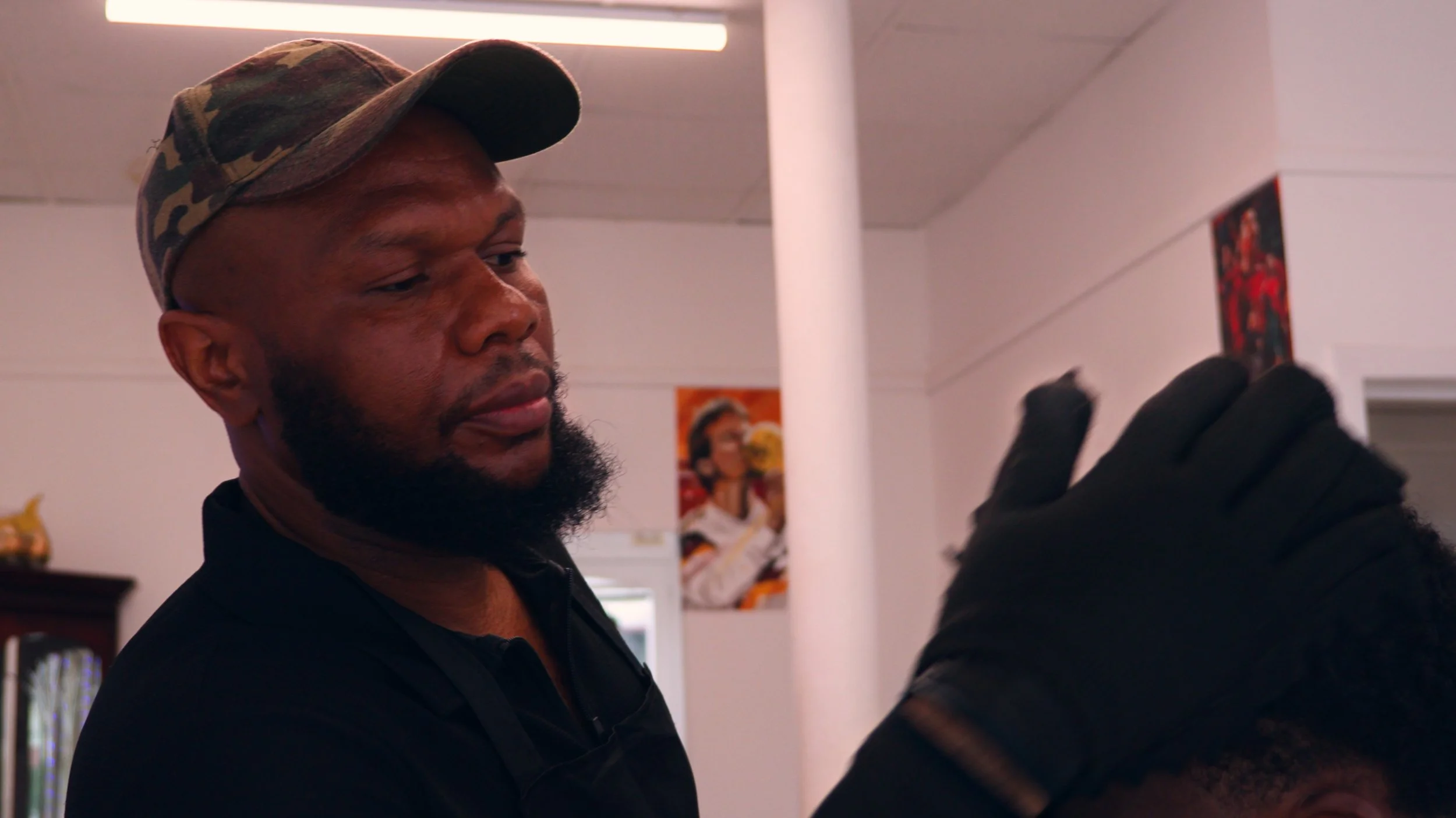 Bearded man wearing a camouflage cap and black shirt sponging up a clients hair.