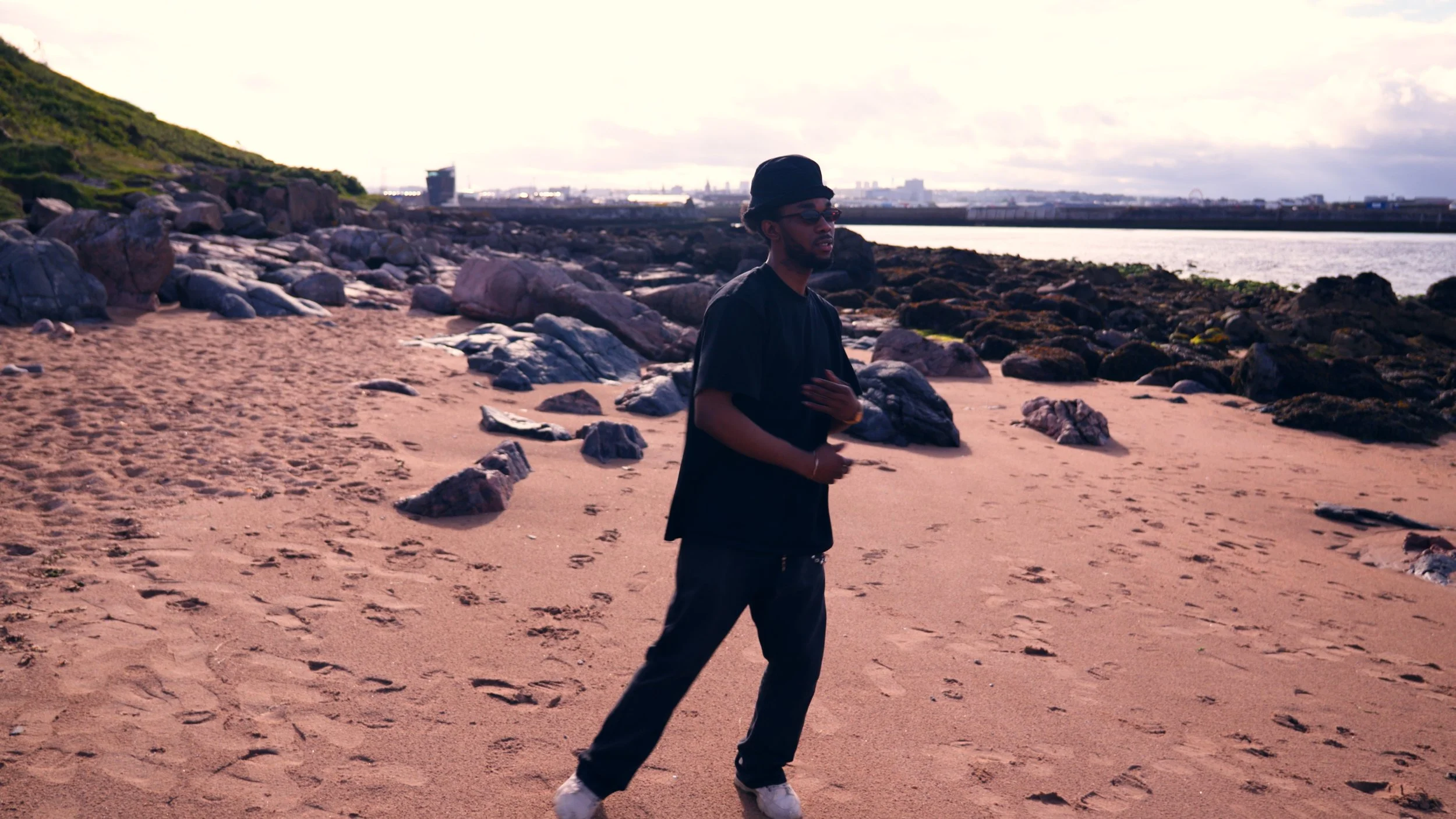 Man in black clothing and bucket hat standing on a rocky beach at sunset, with a coastal city skyline in the background.