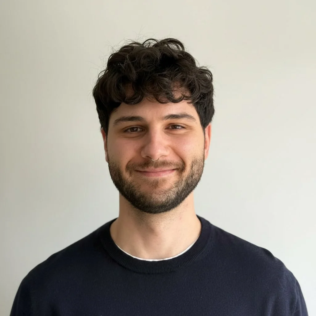 A young man with dark curly hair, a beard, and a light skin tone, smiling softly at the camera, wearing a black shirt, standing against a plain light-colored background.