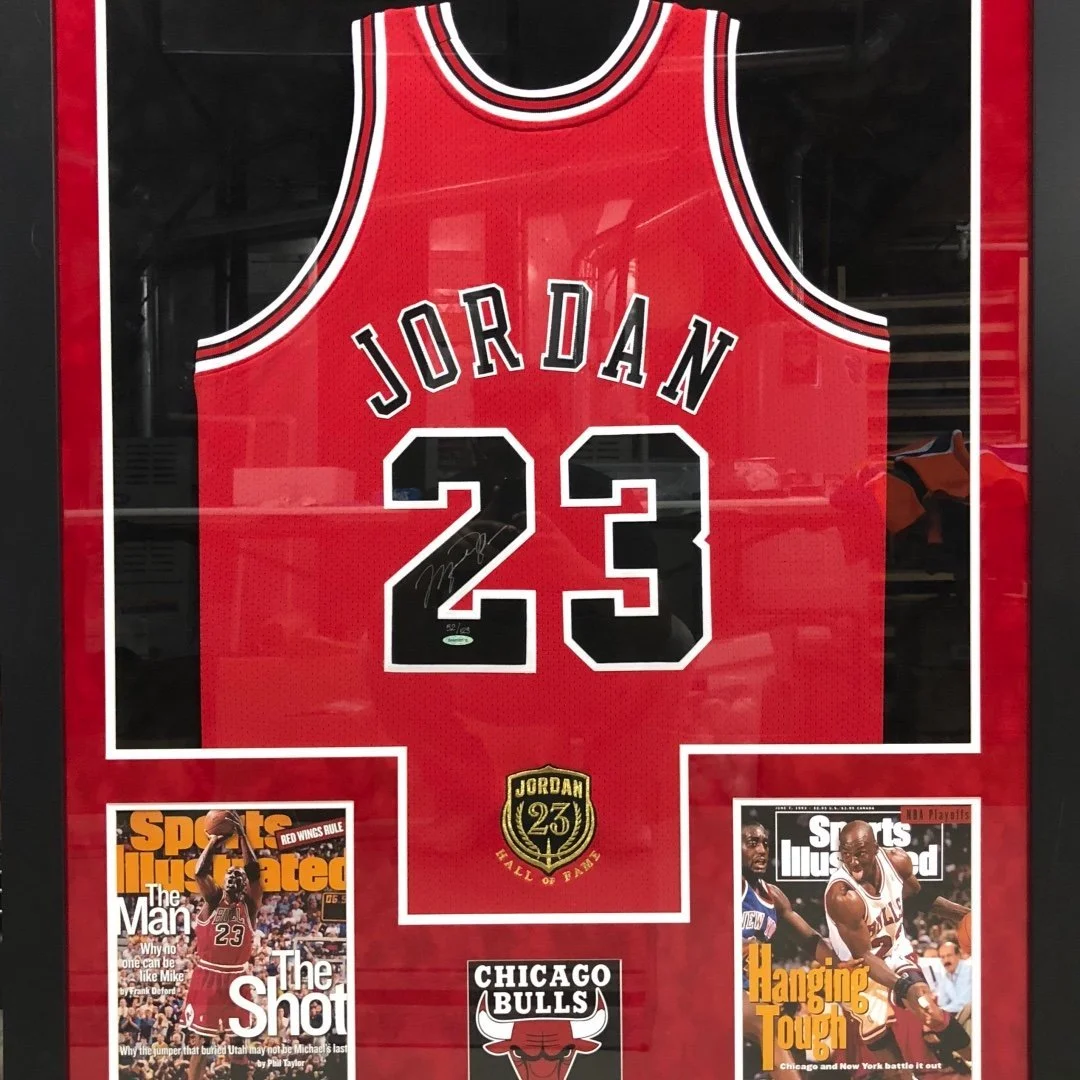 Framed red Chicago Bulls basketball jersey with number 23 and name Jordan, signed on the number, with Sports Illustrated magazine covers featuring Michael Jordan in the bottom corners.