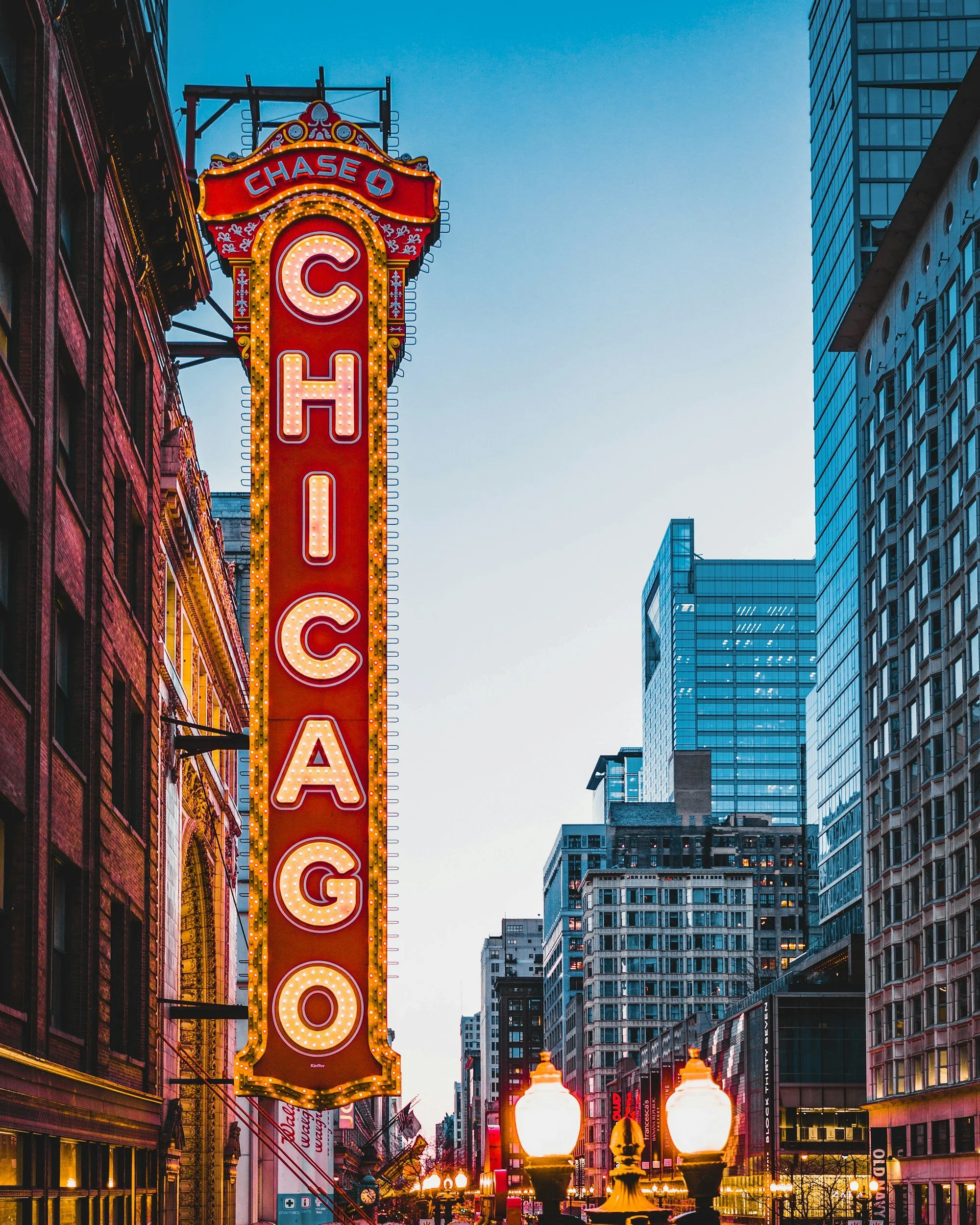 Nighttime view of a city street with tall modern glass buildings and classic brick buildings. A vertical neon sign reads 'CHICAGO' with illuminated bulbs. Street lamps with glowing globes line the street, and various storefronts and signs are visible along the street.