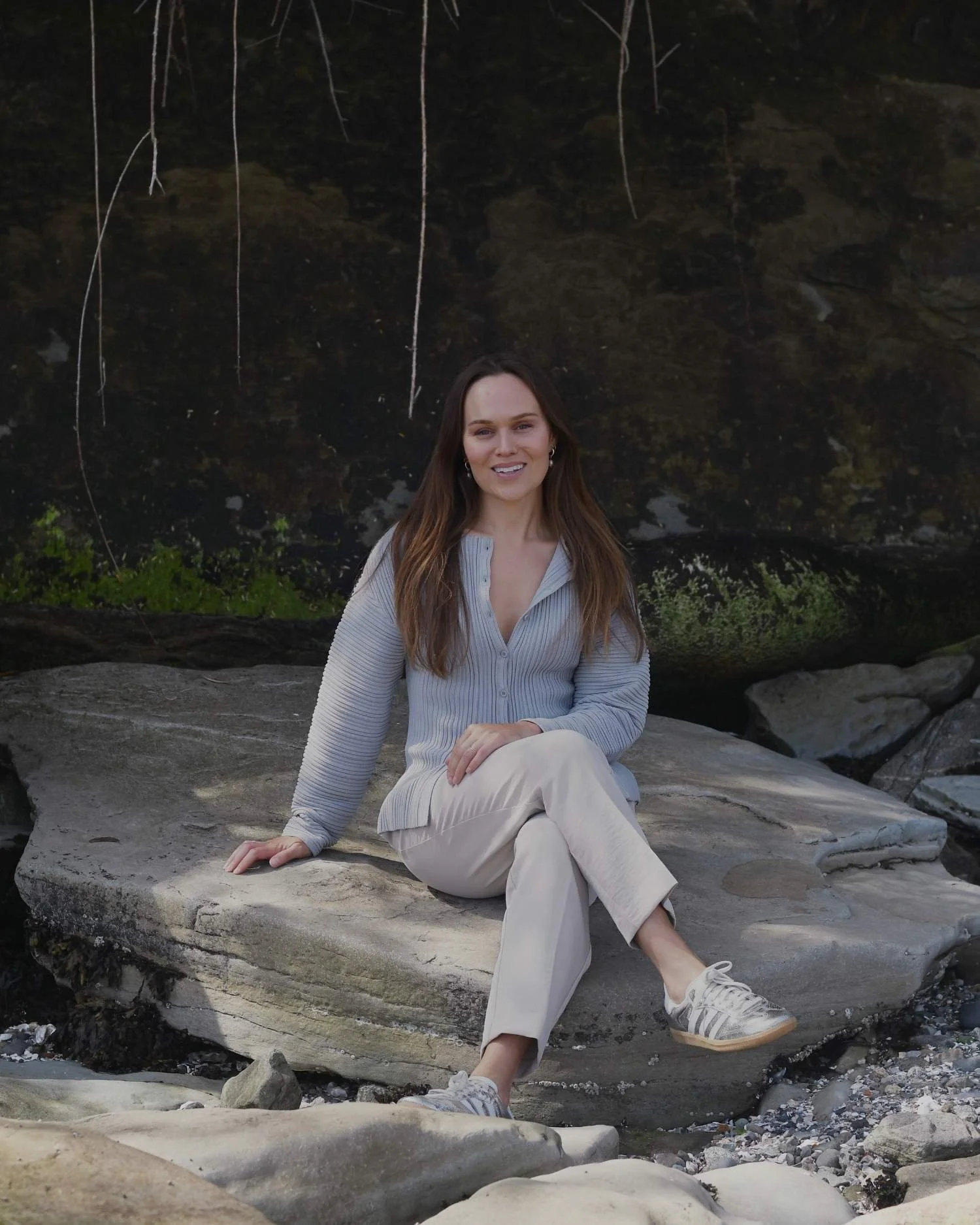 A woman sitting on a large flat rock outdoors, with a dark rocky background and some greenery, smiling and looking at the camera.