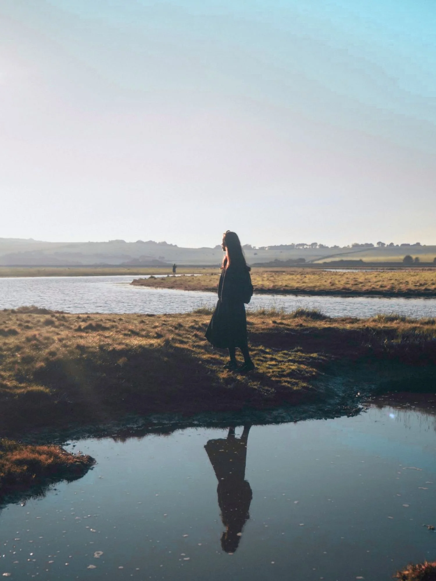 A woman standing by a river with her reflection visible in the water, and a person walking in the background on the other side of the river, with a landscape of rolling hills under a clear sky.