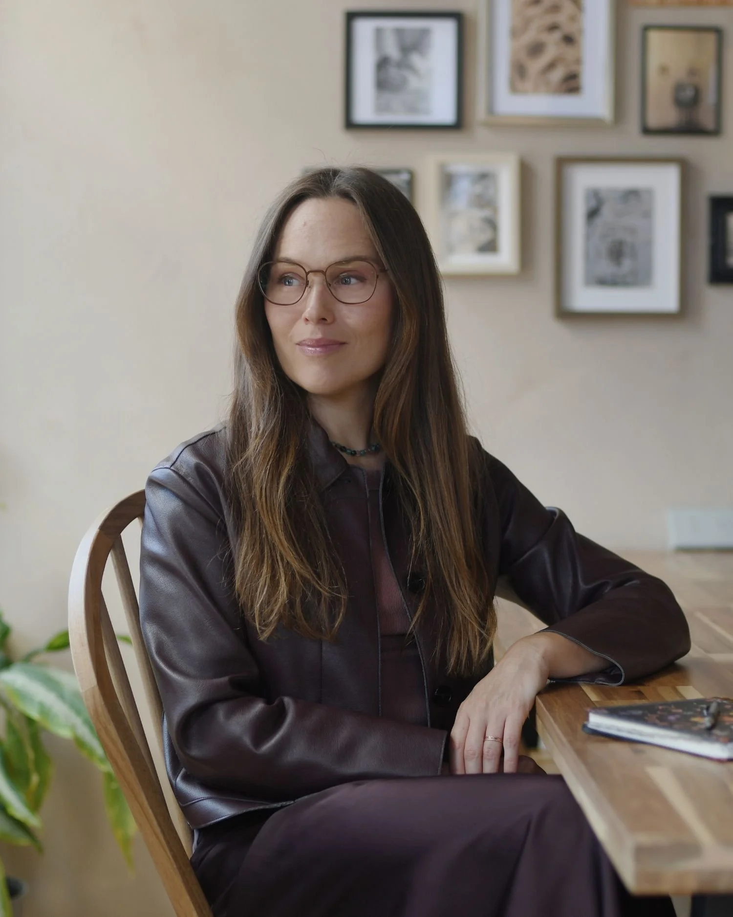 A woman with long brown hair and glasses sitting at a wooden table with a book, in a room decorated with framed pictures on the wall.