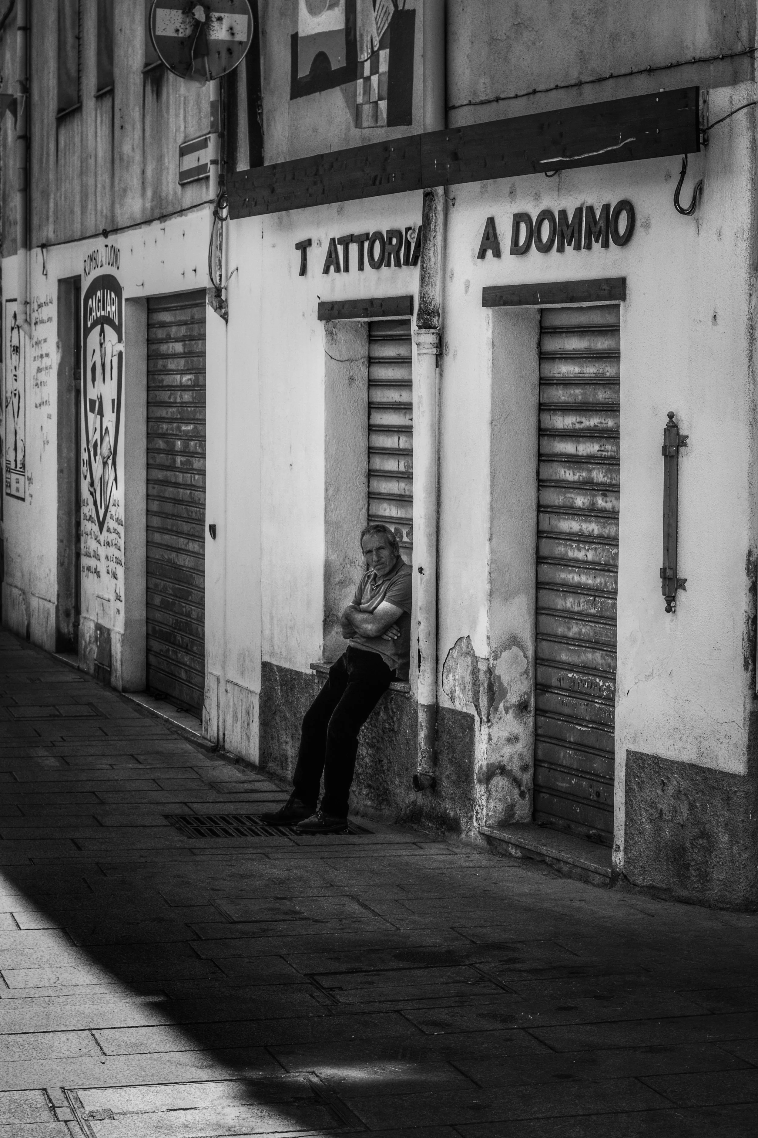 A man leaning against a wall on a city sidewalk with closed shop shutters behind him. The image is in black and white.