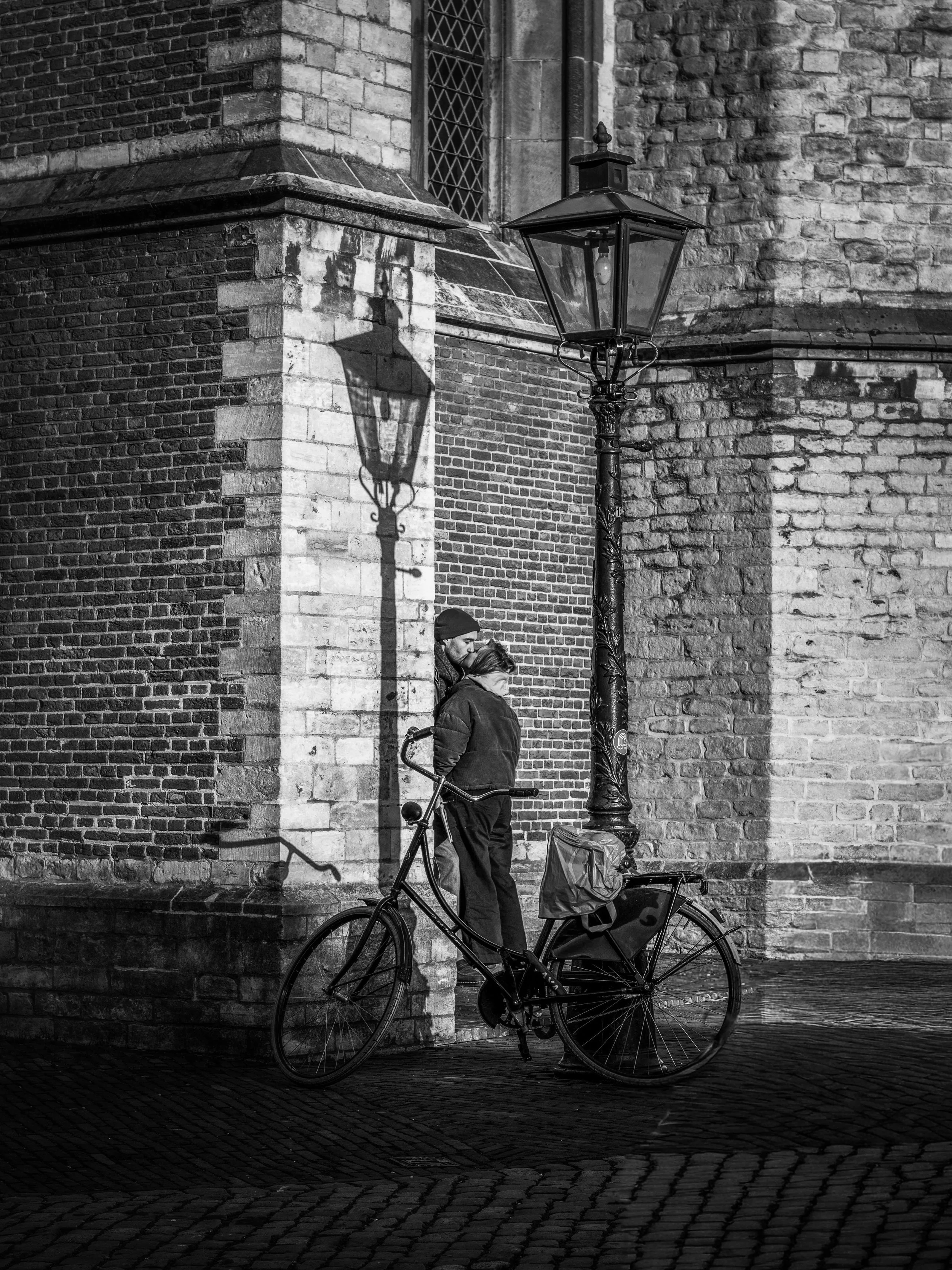 A black and white photo of a street scene at night showing a person with a bike and a bicycle with a cargo compartment, a vintage street lamp, a brick wall, and two people standing close together.