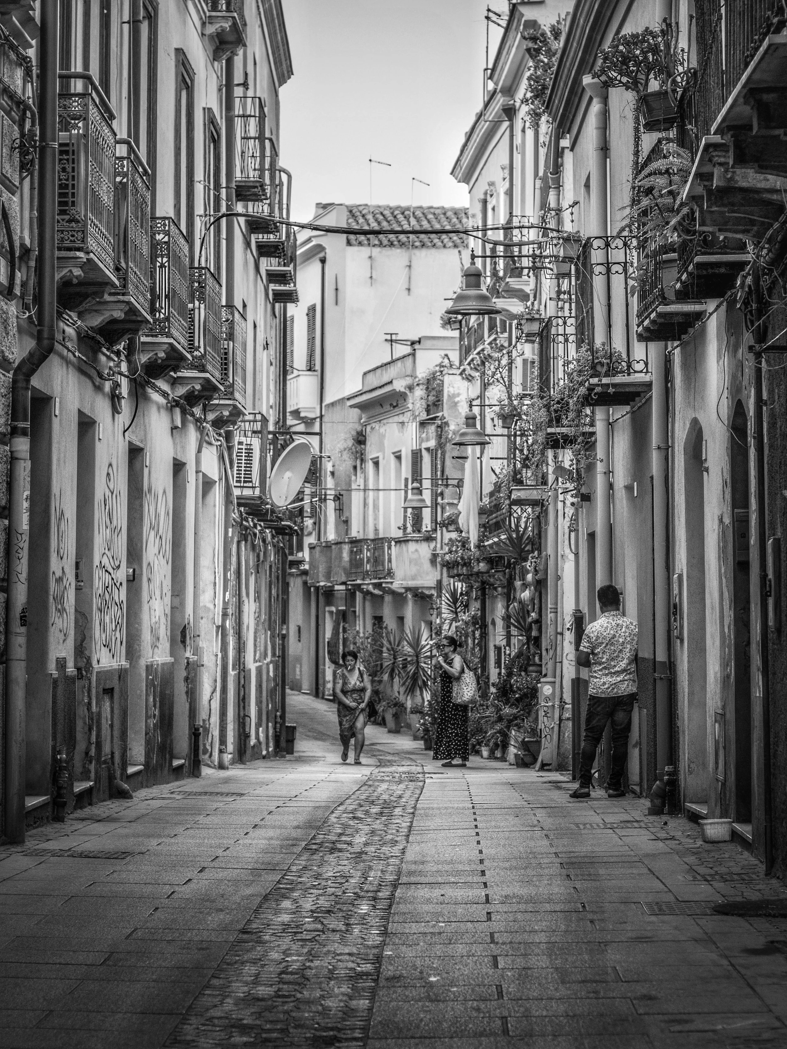 A black-and-white photo of a narrow European street with stone paving, surrounded by old buildings with balconies and potted plants. Several people are walking and standing on the street.